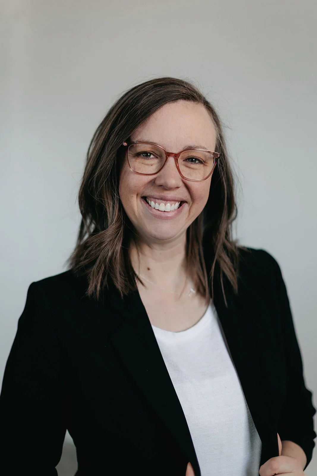 A woman with shoulder-length brown hair, wearing glasses, a white shirt, and a black blazer, smiling at the camera against a plain, light-colored background.