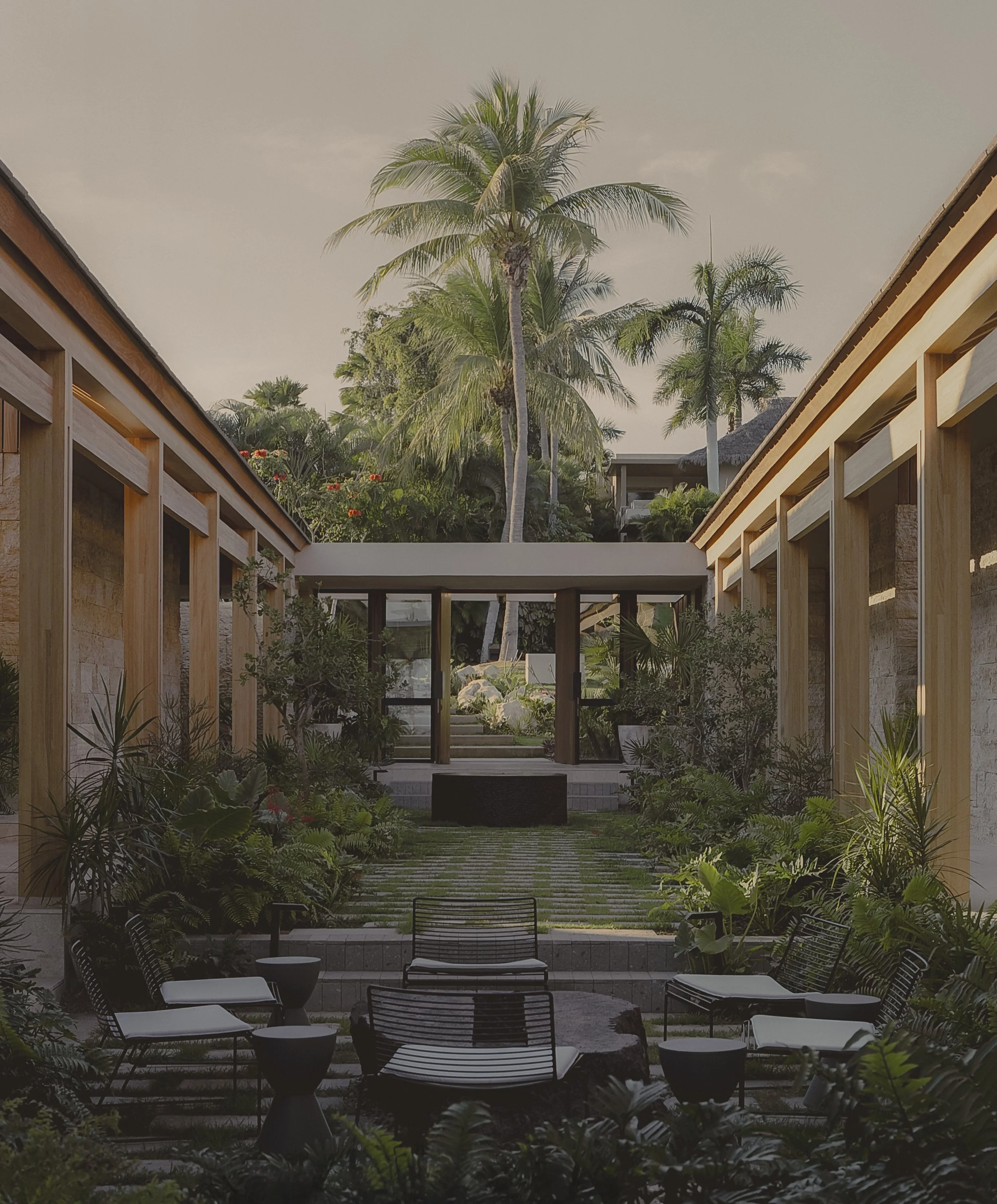 Arquitectura moderna con paredes de piedra y madera, entrada con puertas abiertas, rodeada de plantas verdes y rocas decorativas.