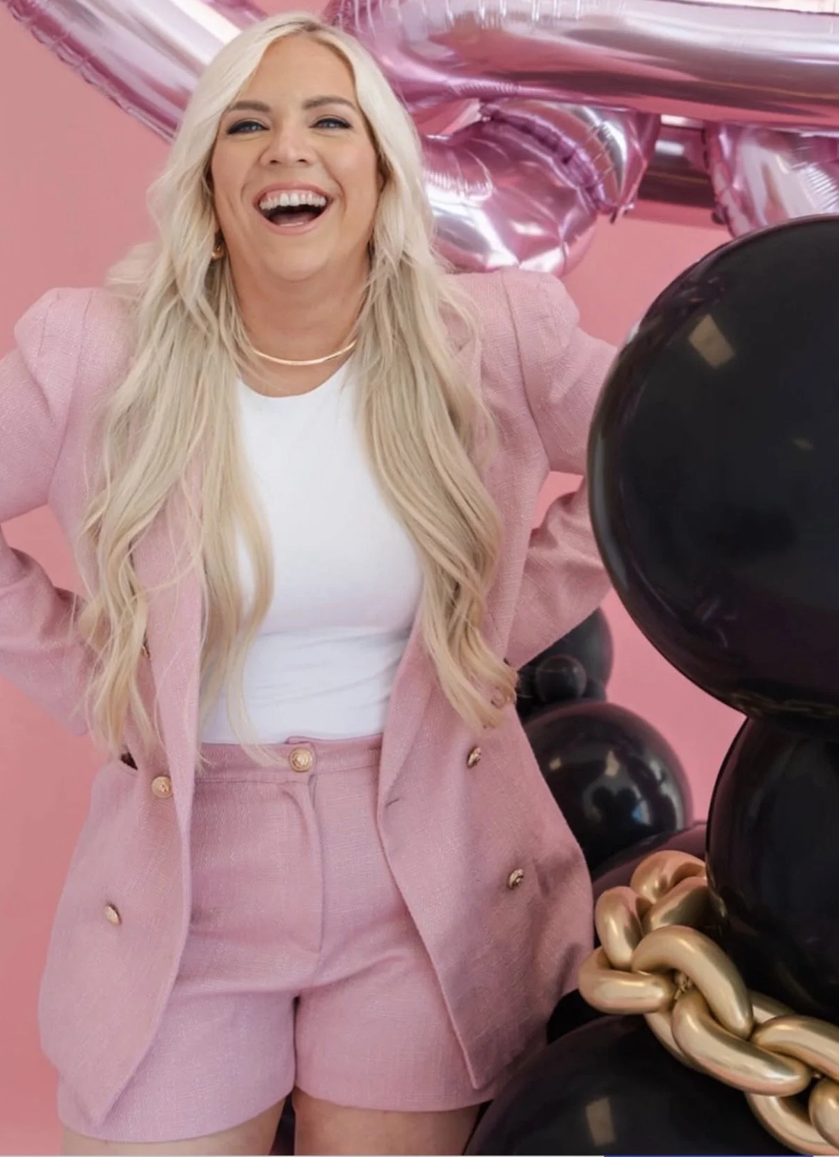 A woman in a pink blazer and pink shorts with gold buttons, wearing a white top and gold necklace, standing in front of a pink background with metallic pink balloons and black, gold, and pearl-colored balloon sculptures.