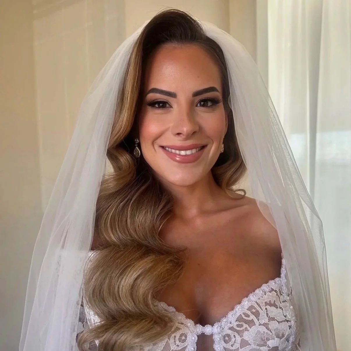 A smiling woman in a white wedding dress with a veil and earrings, standing indoors near a window.