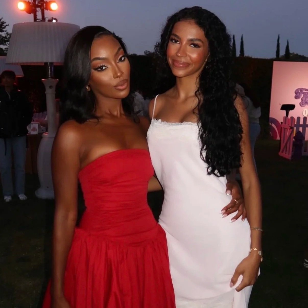 Two women standing close together at an outdoor event during dusk. The woman on the left is wearing a strapless red dress, and the woman on the right is wearing a white dress with spaghetti straps. They are smiling and have their arms around each oth