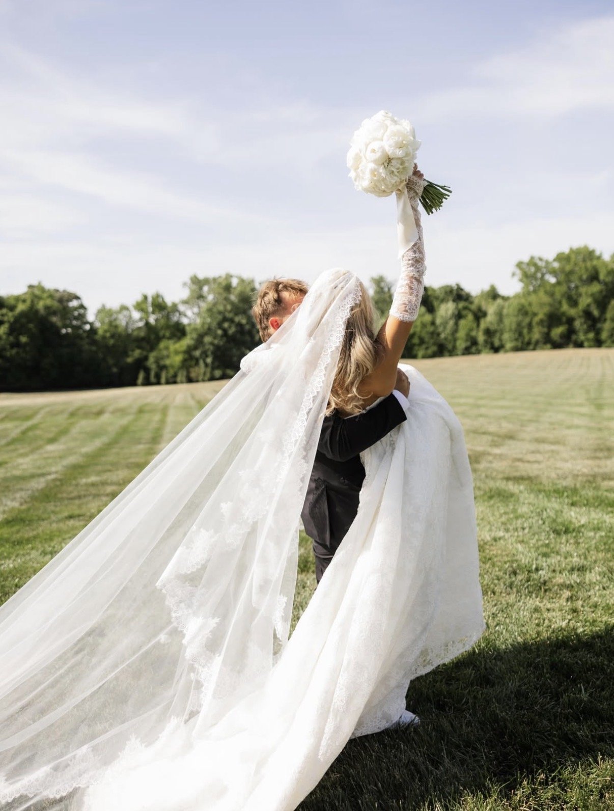 A bride in a white wedding dress with lace veil holding a bouquet of white flowers, being lifted by a groom in a black tuxedo across a grassy field with trees in the background on a sunny day.