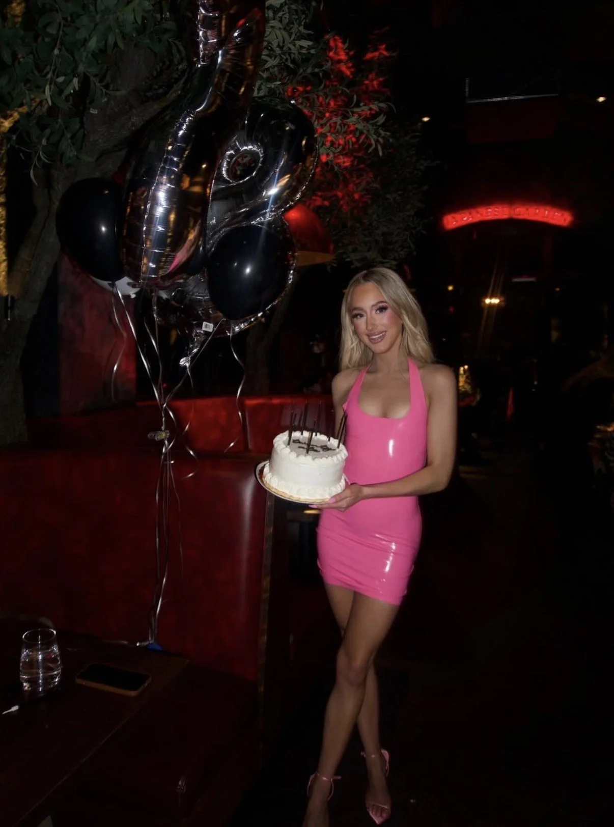 A woman in a pink dress holding a birthday cake with candles, standing next to black balloons in a dimly lit setting with red lighting and greenery in the background.