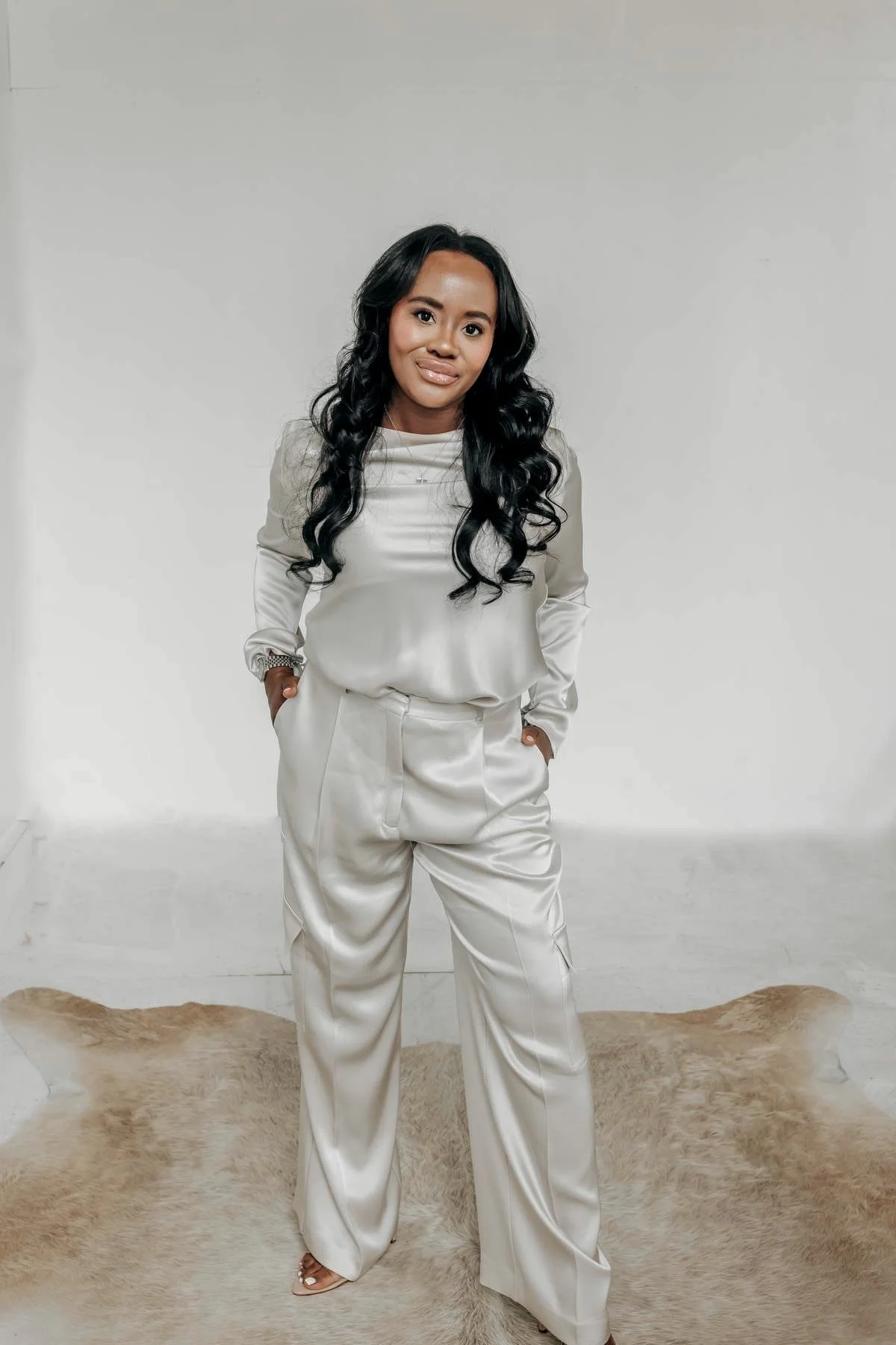 A woman with long black curly hair, wearing a shiny beige satin blouse and matching satin pants, standing on a beige fur rug in front of a plain white wall.