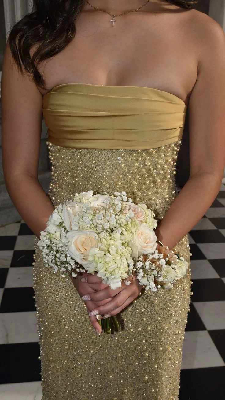 A woman in a strapless, gold, beaded gown holding a bouquet of white and blush roses with baby's breath flowers in an elegant setting.