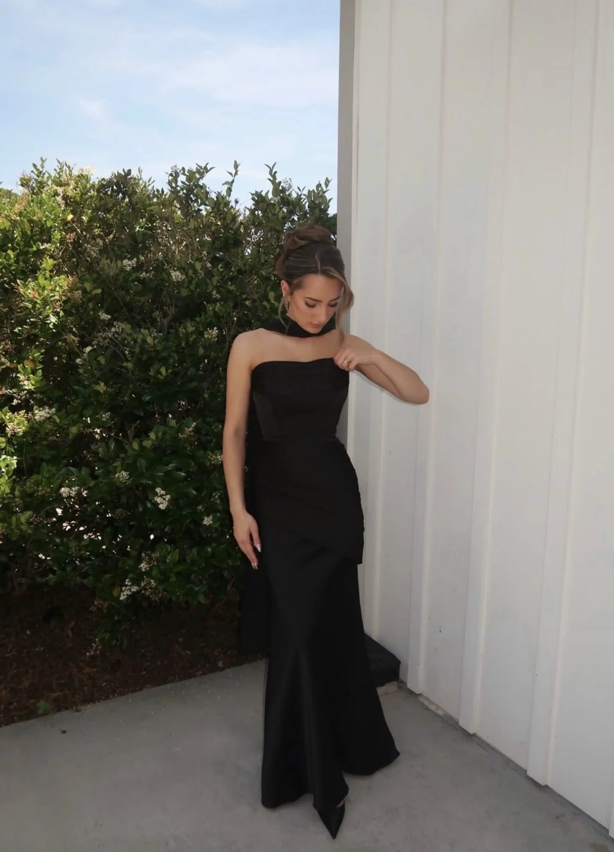 Woman in a black strapless evening gown standing outdoors next to a white wall with bushes in the background.