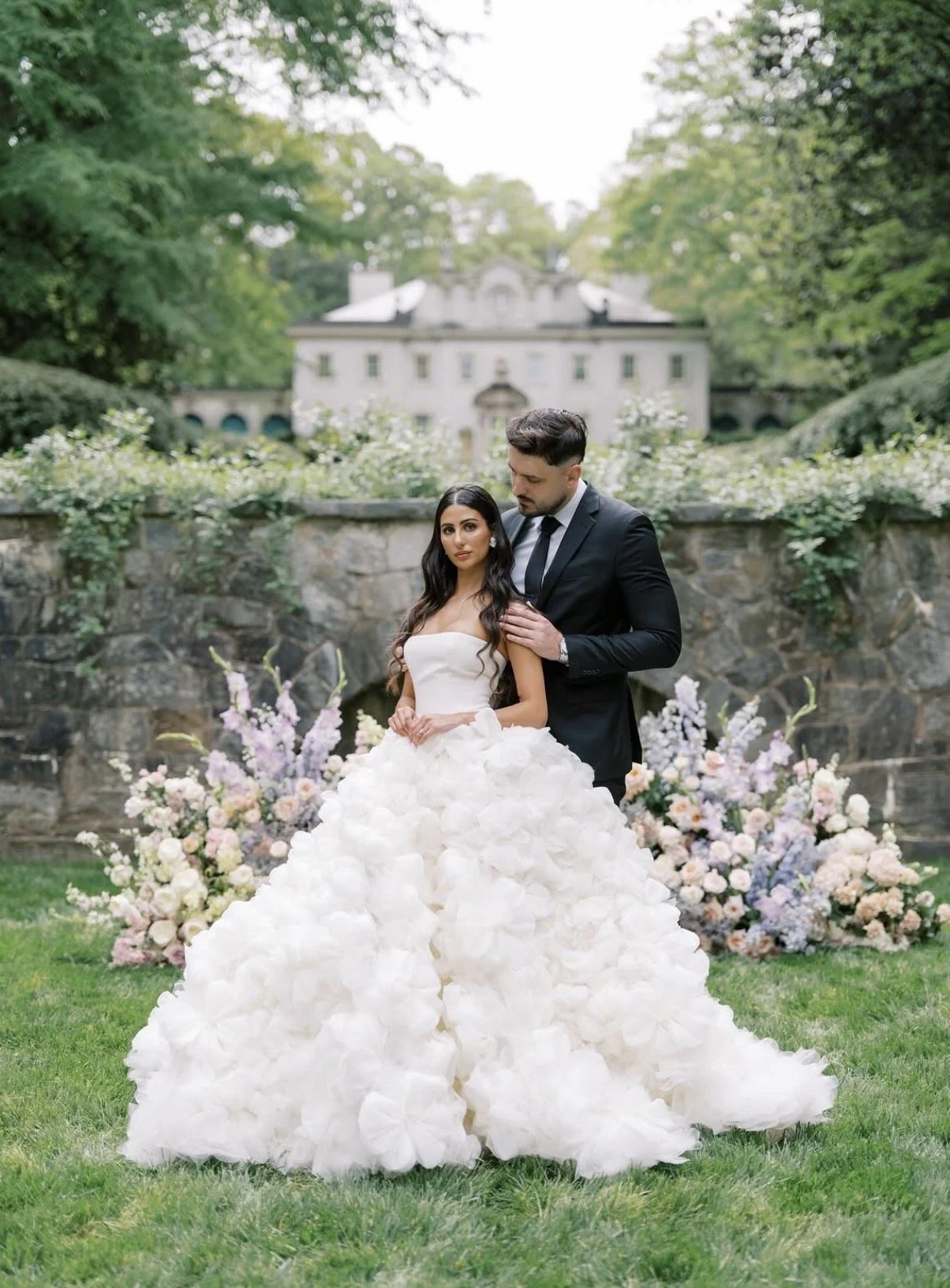 A bride and groom standing outdoors on a wedding day, with the bride in a white gown with a voluminous skirt, and the groom in a black suit with a tie. They are surrounded by a floral arrangement and greenery, with a historic building in the background.
