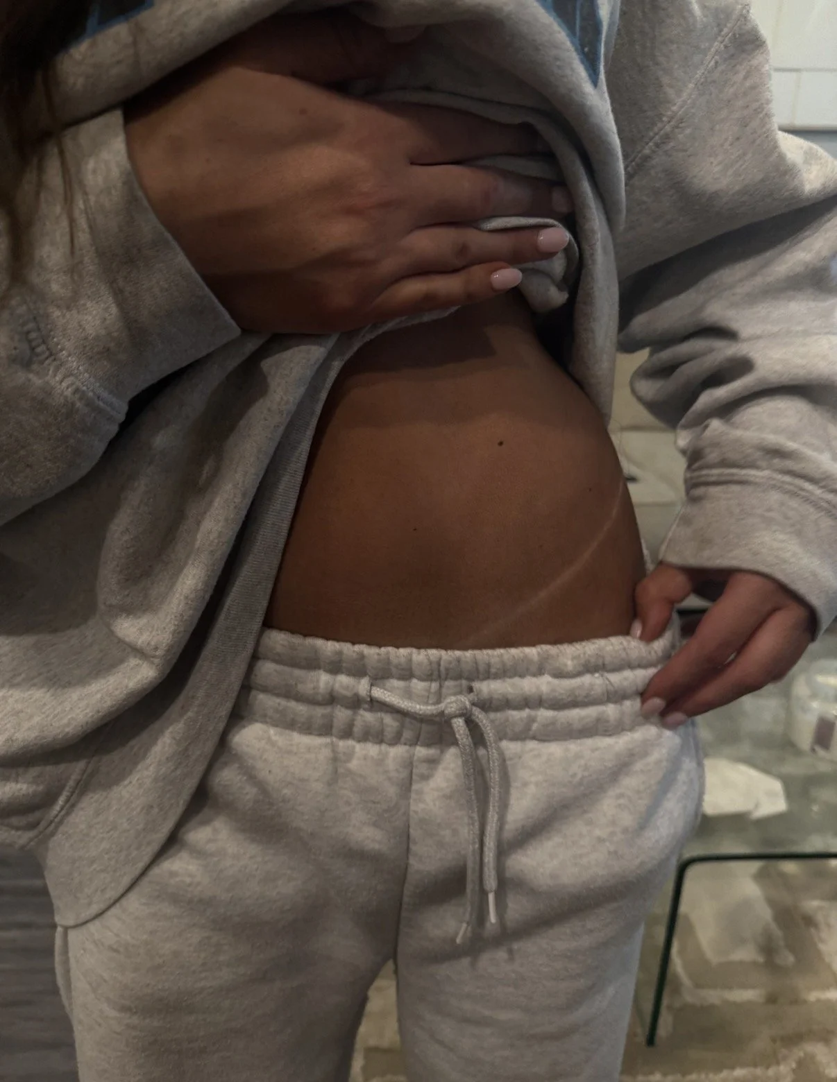 Person wearing gray sweatpants and a matching hoodie, lifting the hoodie to reveal their midsection. The person has light skin and neatly manicured nails. The background appears to be indoors with a tiled wall and some furniture.
