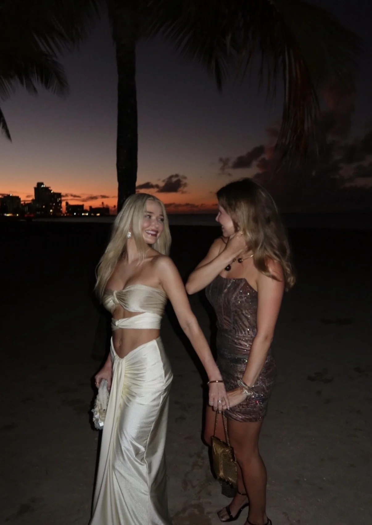 Two women standing on the beach at sunset, holding hands and smiling at each other, with palm trees and buildings in the background.
