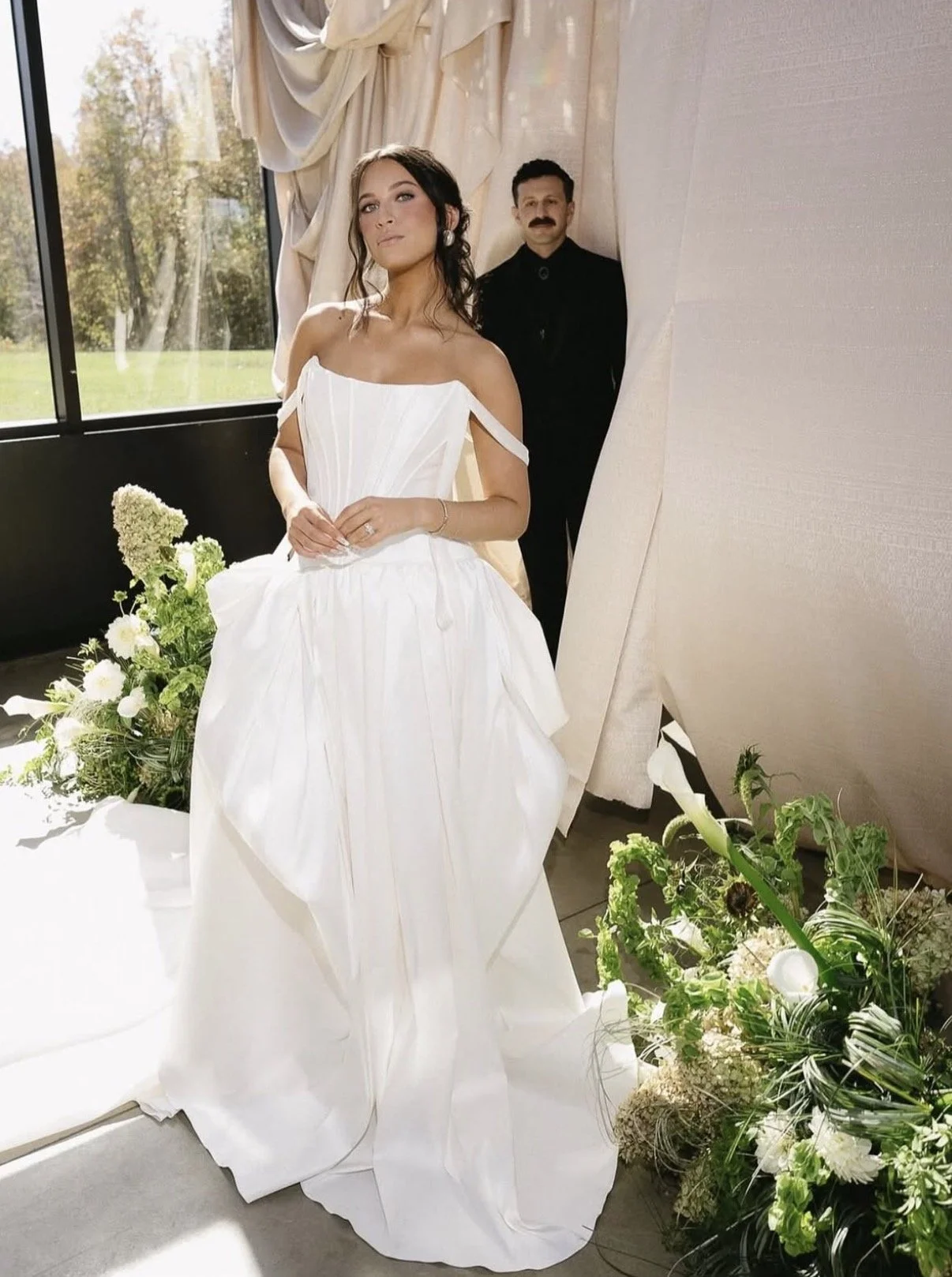 A woman in a white wedding dress standing next to a large bouquet of white and green flowers, with a man in a black suit standing behind her. They are inside a room with large windows and cream-colored drapes.