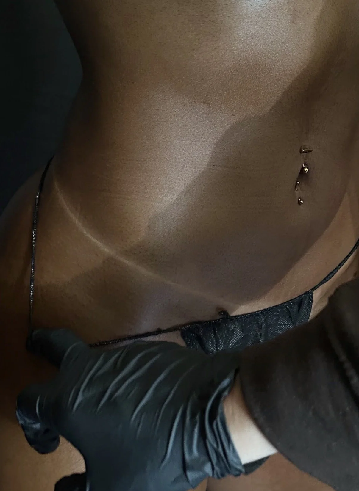 Close-up of a person's chest with nipple piercings, wearing a black lace bra, and a person in striped shorts leaning over.