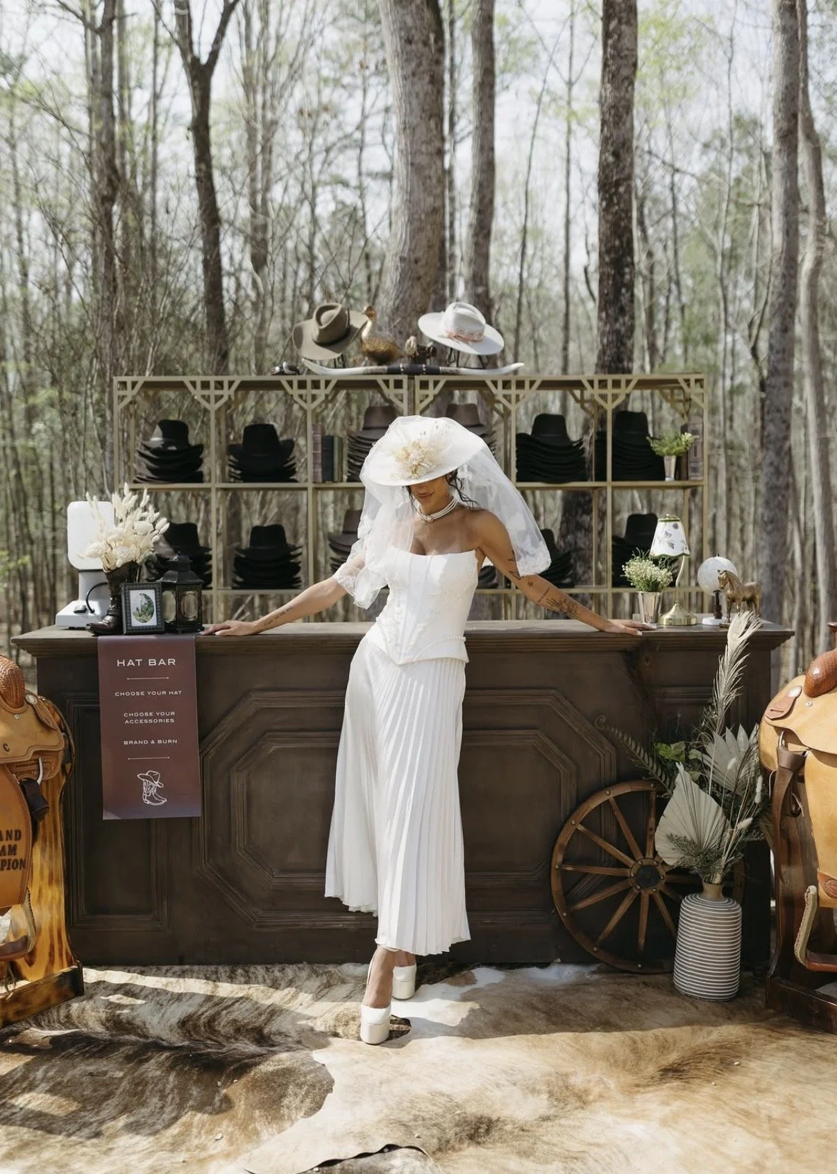 A woman dressed in a white bridal gown and veil with a large hat stands at a rustic outdoor hat bar surrounded by wilderness trees. The bar has shelves with black hats, decorative items, and flowers. The setting is decorated with vintage accessories, horses, and greenery, creating a vintage western wedding theme.