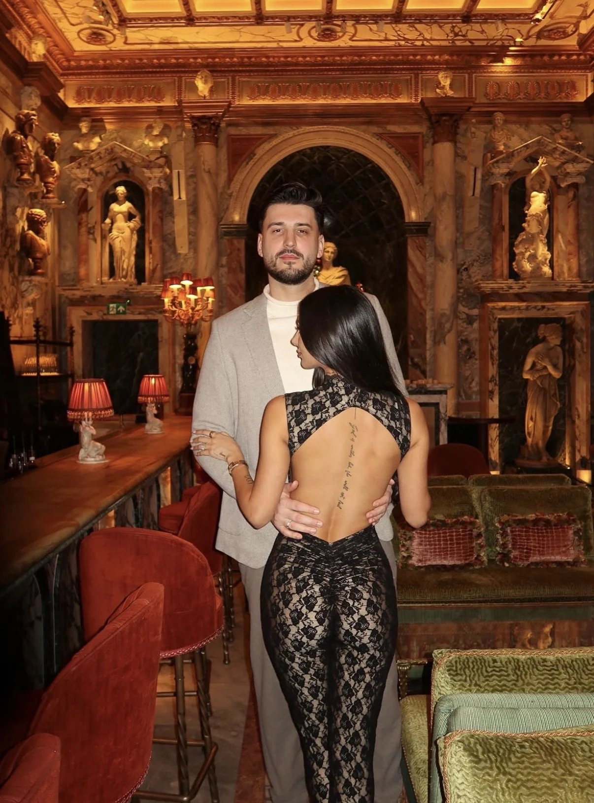 A man and woman in an ornate, historic-looking room with sculptures and gold accents. The woman is wearing a revealing black lace dress with an open back and a tattoo on her spine. The man is dressed in a light-colored suit and stands beside her with