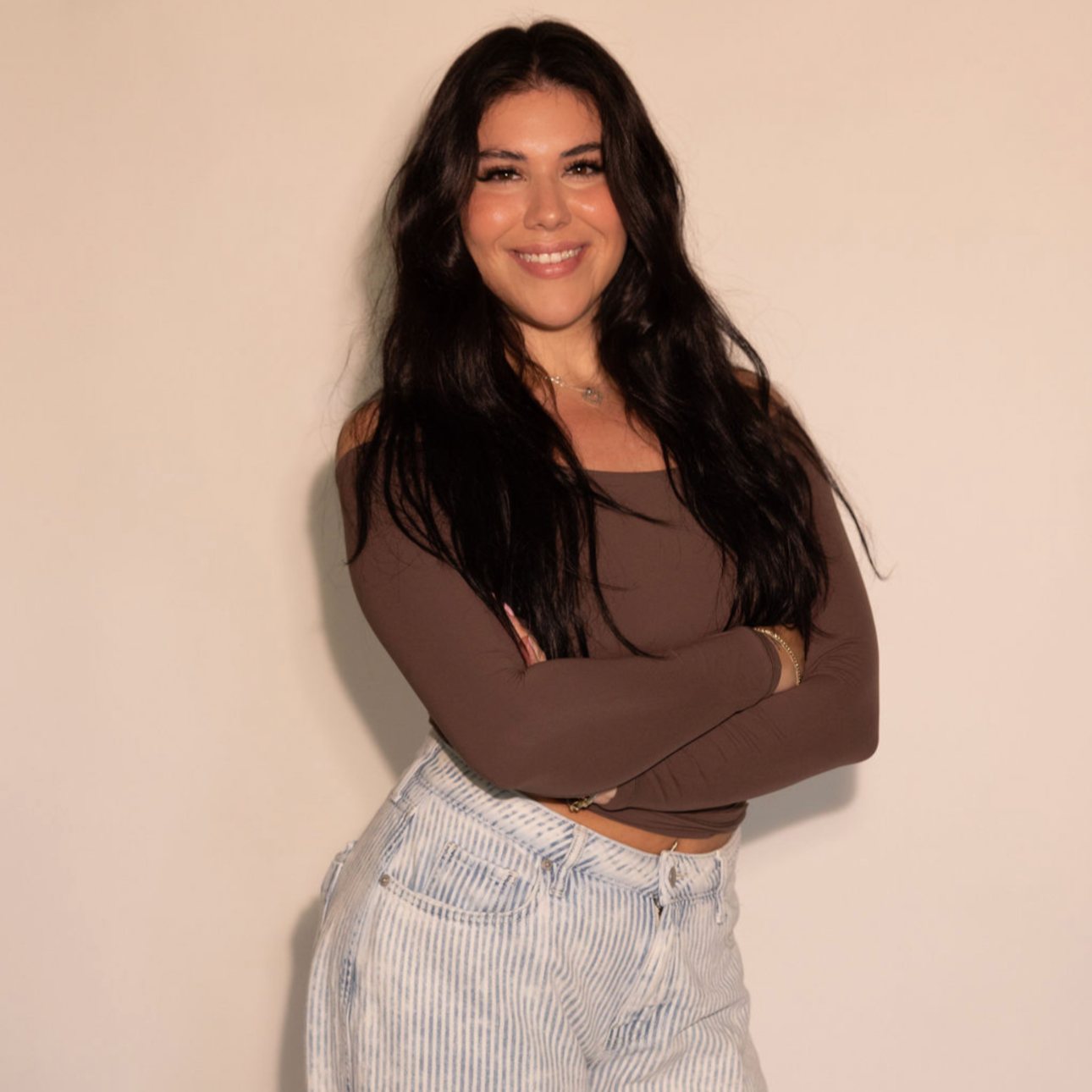 A young woman with long dark hair smiling, arms crossed, wearing a brown long-sleeve top and high-waisted striped pants, standing against a plain off-white wall.