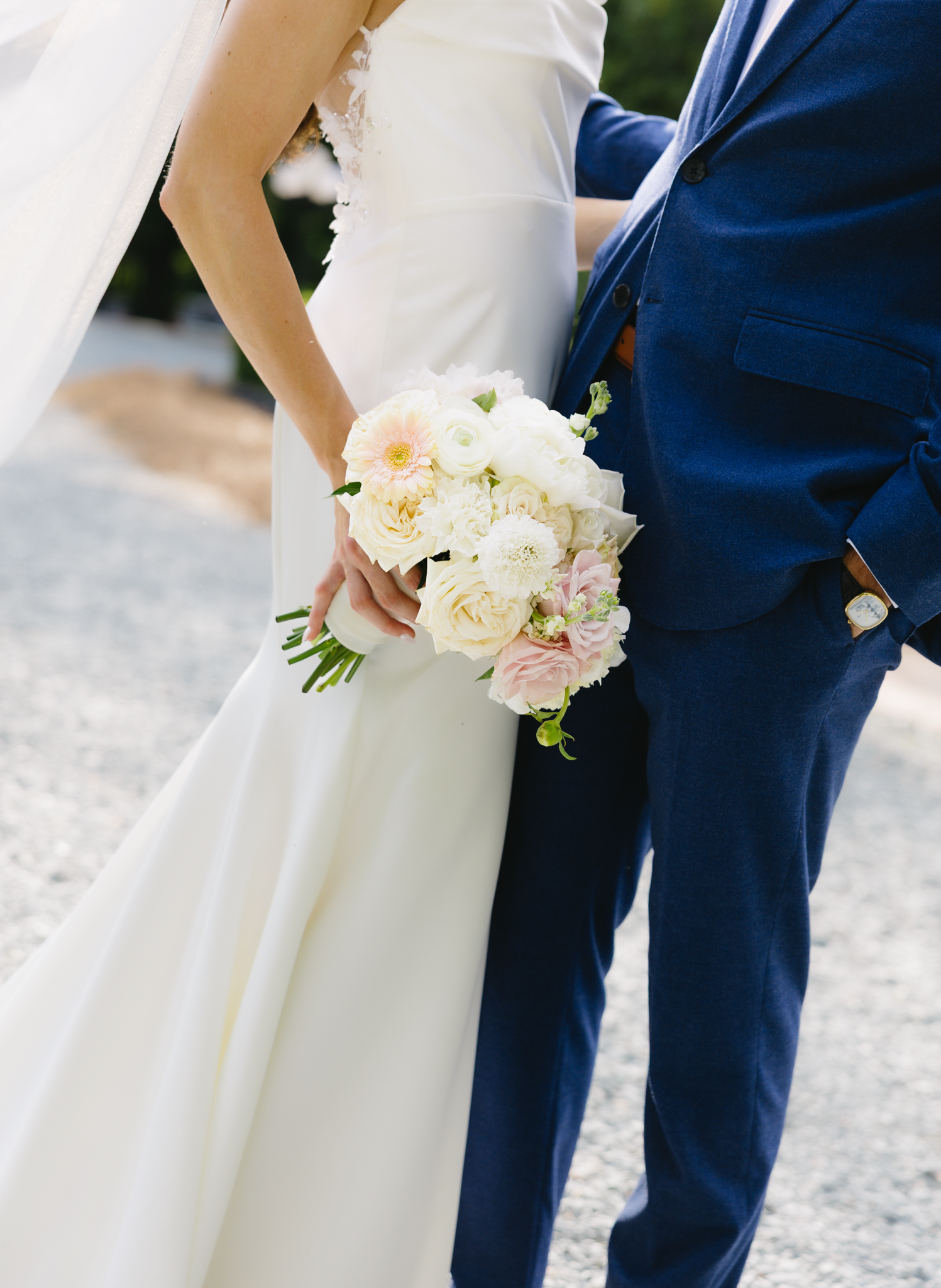 Close-up of a bride and groom standing outdoors. The bride is holding a bouquet of white and pale pink flowers. She is wearing a white wedding dress, and the groom is wearing a blue suit. The photo captures the moment they are holding hands.