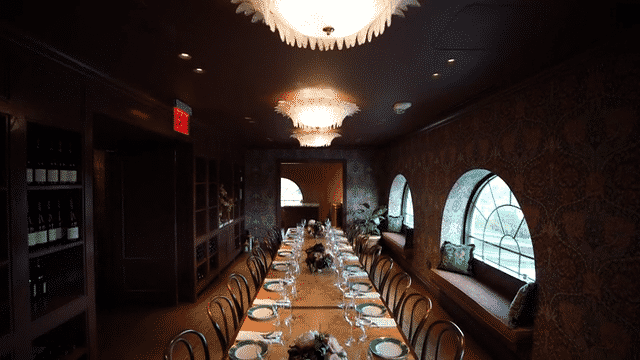 Long dining table set with plates, glasses, and napkins in a dimly lit, elegant restaurant with arched windows and decorative wallpaper.