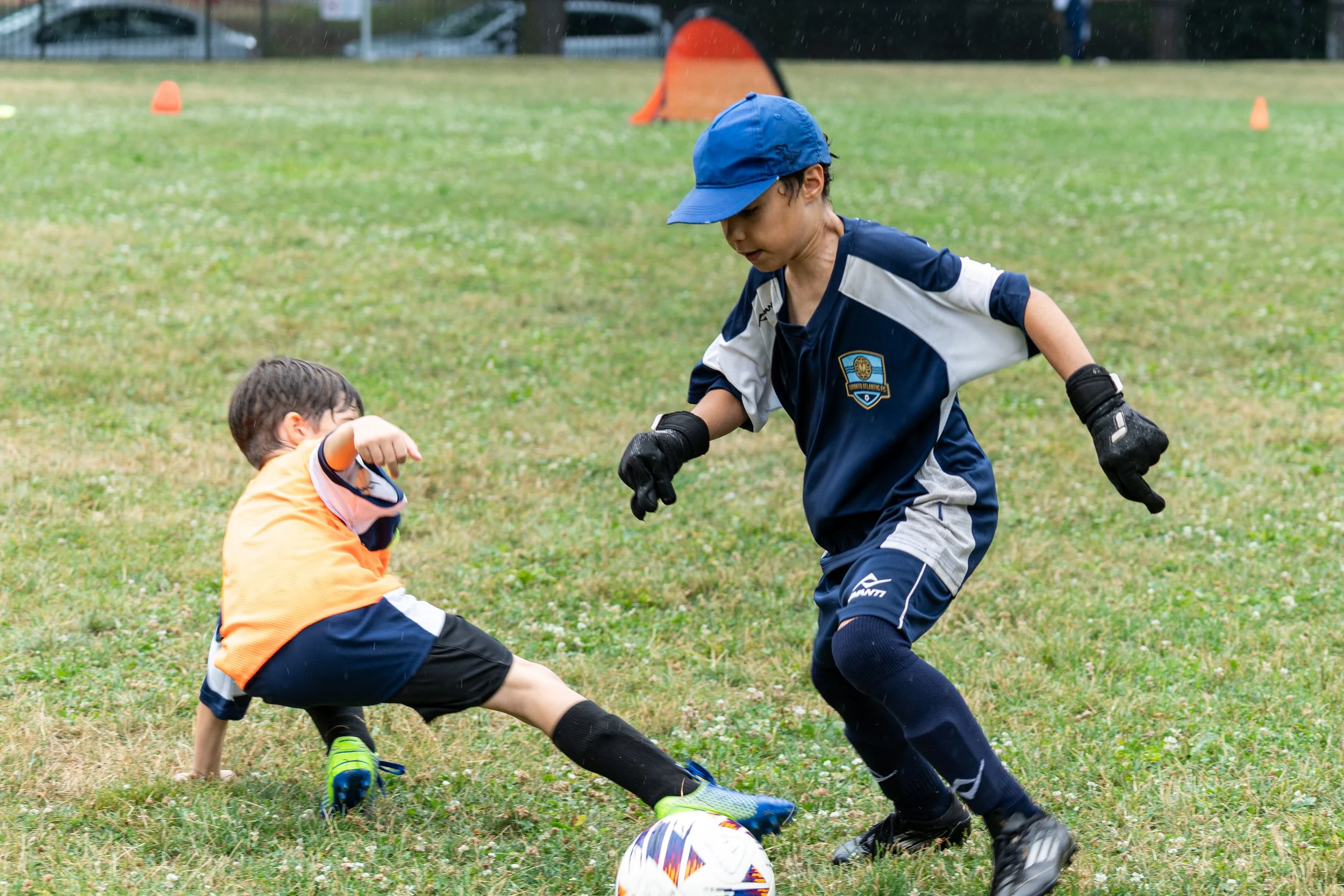 A happy young soccer player balancing a ball on his head while smiling, with an attentive teammate beside him during training at Toronto Atlantic FC, a youth soccer club in Toronto.