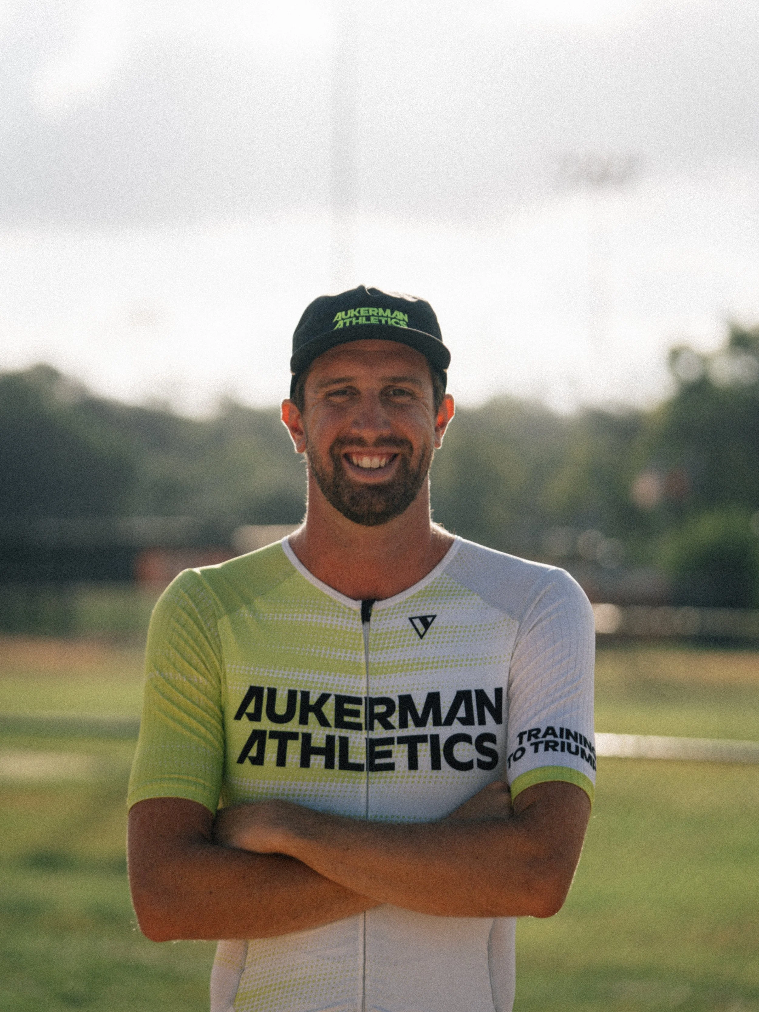 A man in athletic clothing and a cap, smiling with arms crossed, standing outdoors on a sunny day.