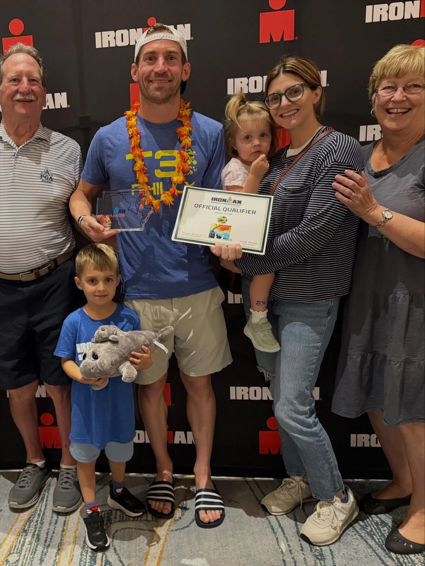 Group of six people, including a child holding a plush animal, posing at an Ironman event with a black backdrop displaying the Ironman logo; one man wears a leis and holds an award, another woman holds a young girl, and others smile.