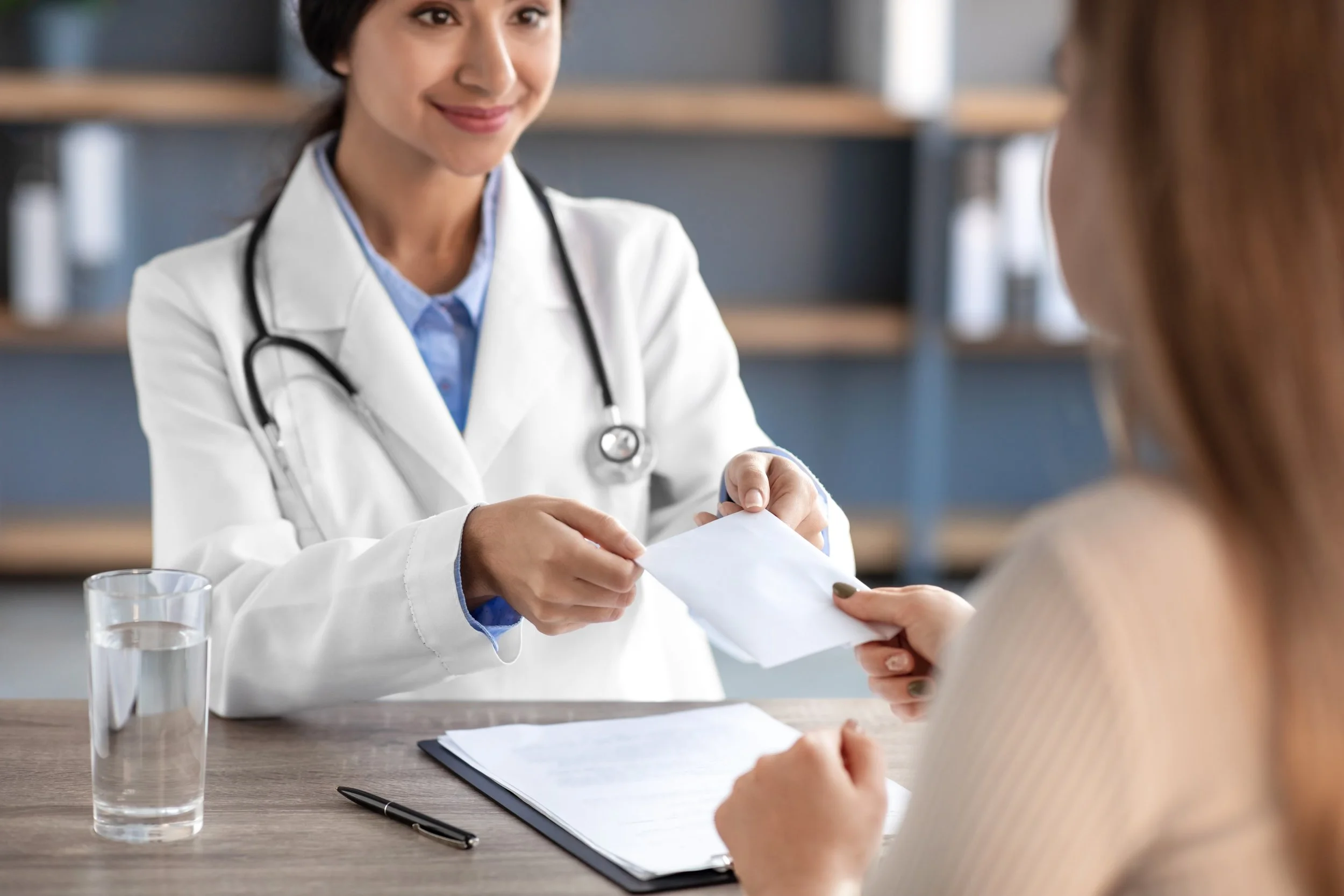 A female doctor in a white coat with a stethoscope around her neck handing a prescription to a patient in a beige sweater. They are sitting at a wooden table in a medical office, with a glass of water, a document, and a pen on the table.