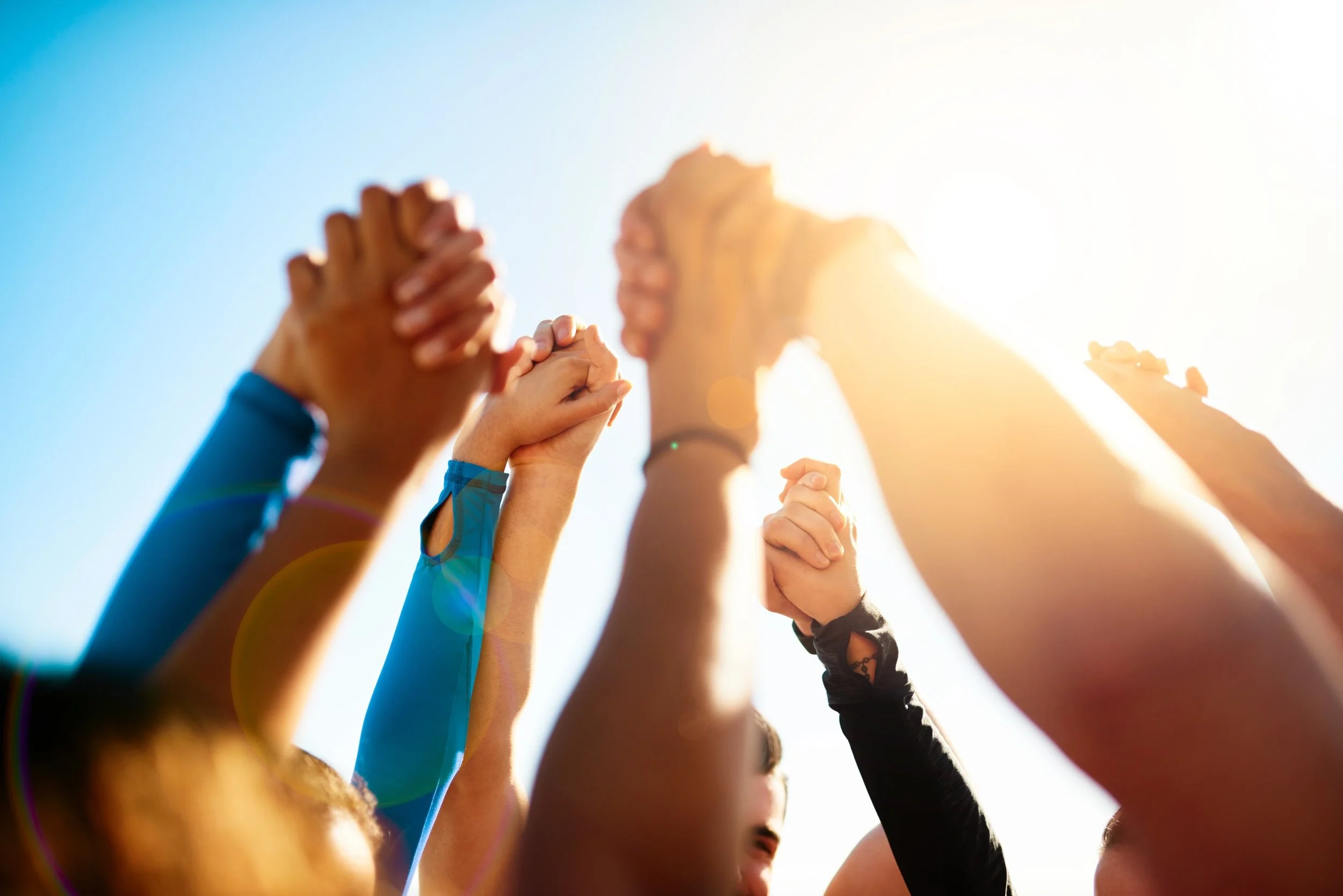 Group of people raising their hands together in a circle outdoors with bright sunlight.