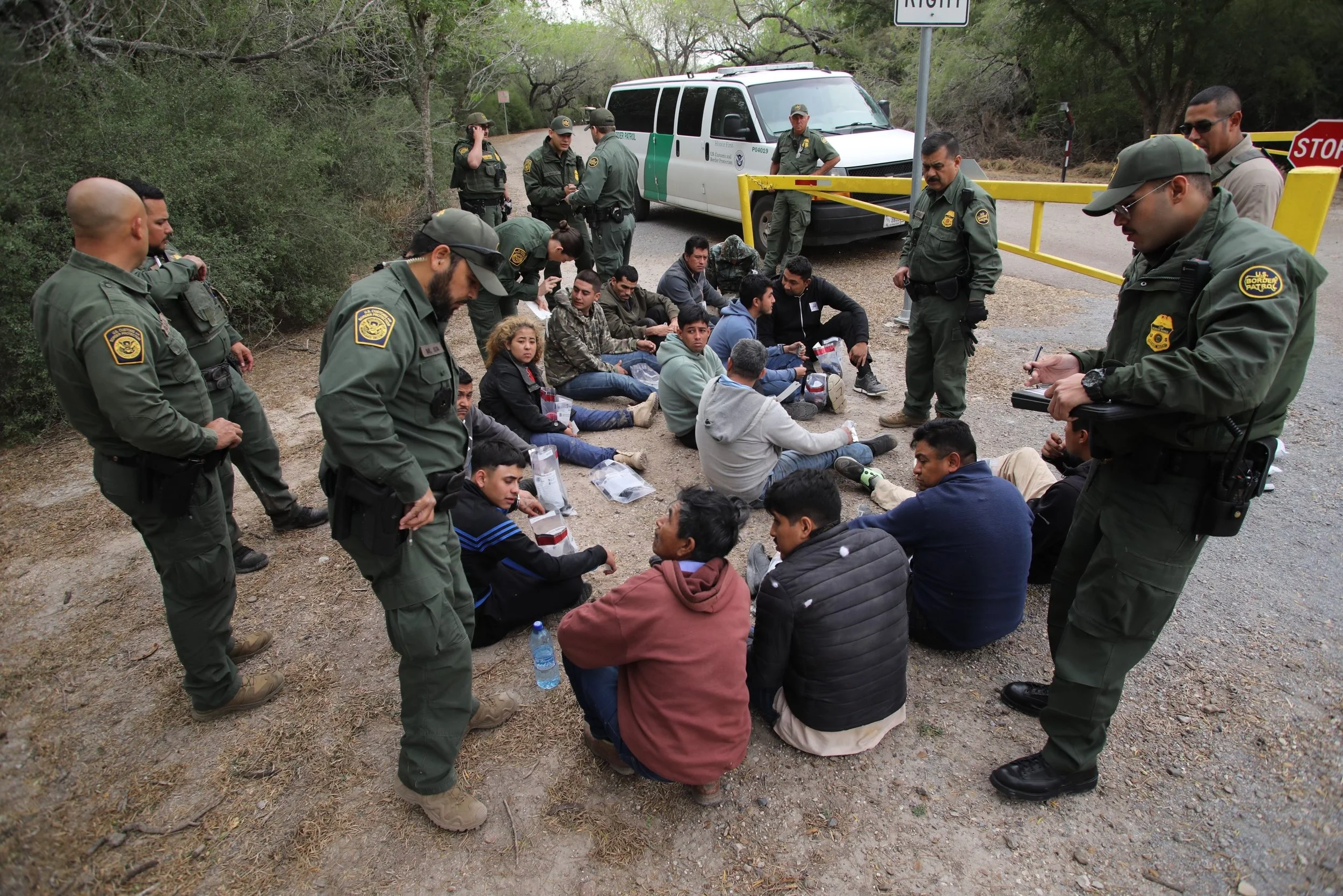 Border patrol agents are overseeing a group of migrants sitting on the ground by a locked gate in a rural area.