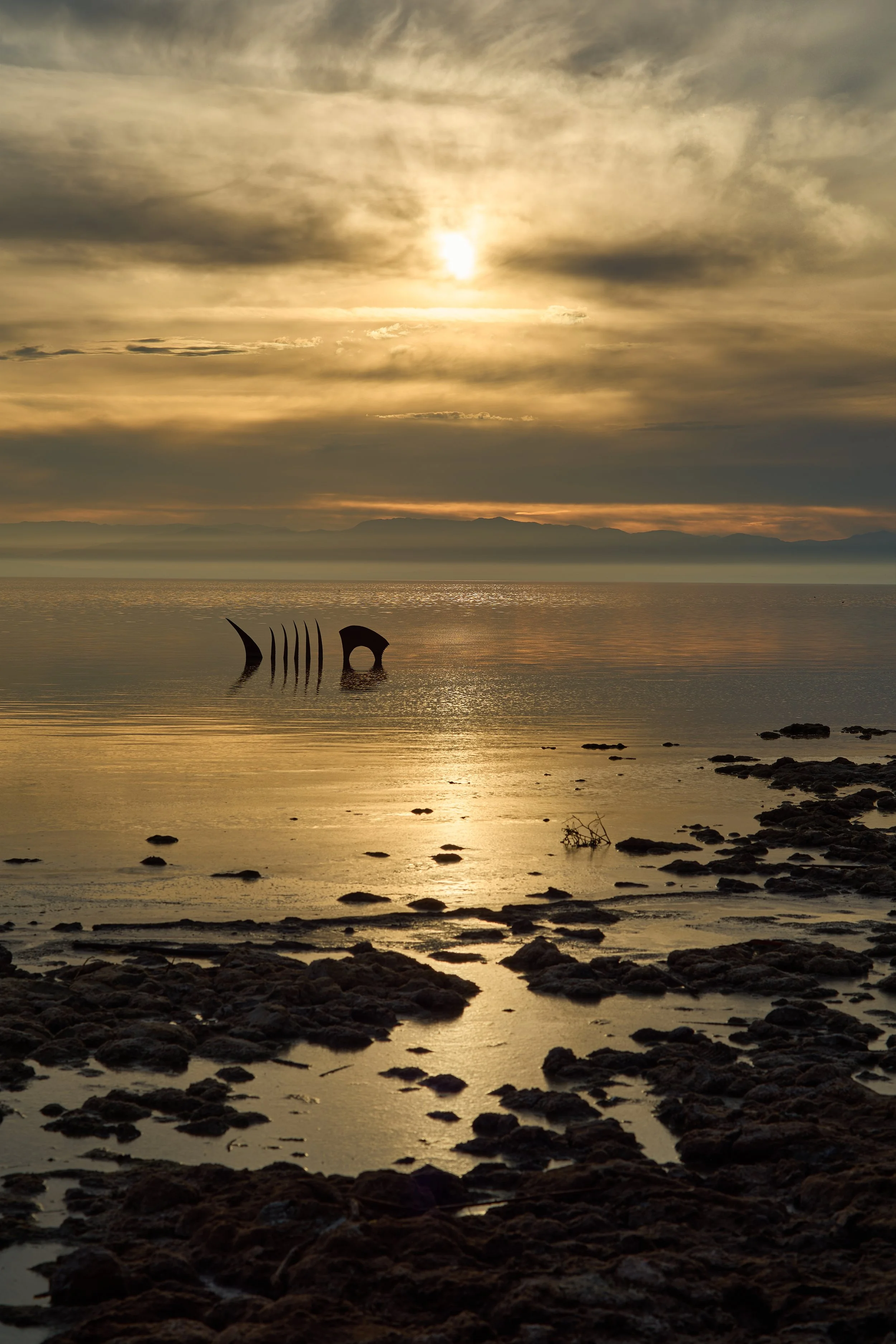 Sunset over a body of water with distant mountains in the background, displaying a sky with golden and gray clouds, and the silhouette of a large metal structure partially submerged in the water near the shoreline. Bombay Beach, Salton Sea, CA.
