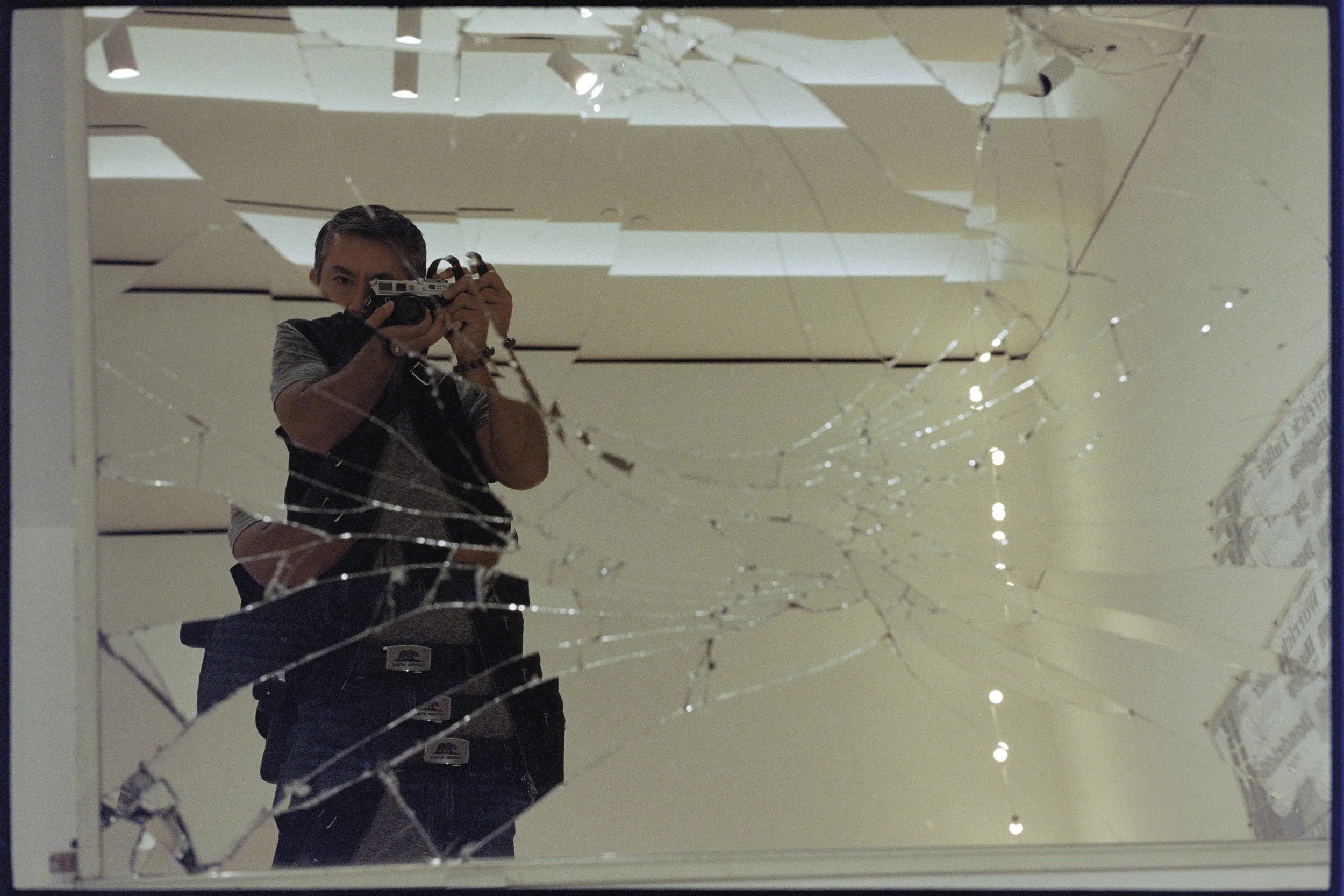 A person taking a photo of their reflection in a shattered mirror, with the beam of the ceiling lights visible in the background.