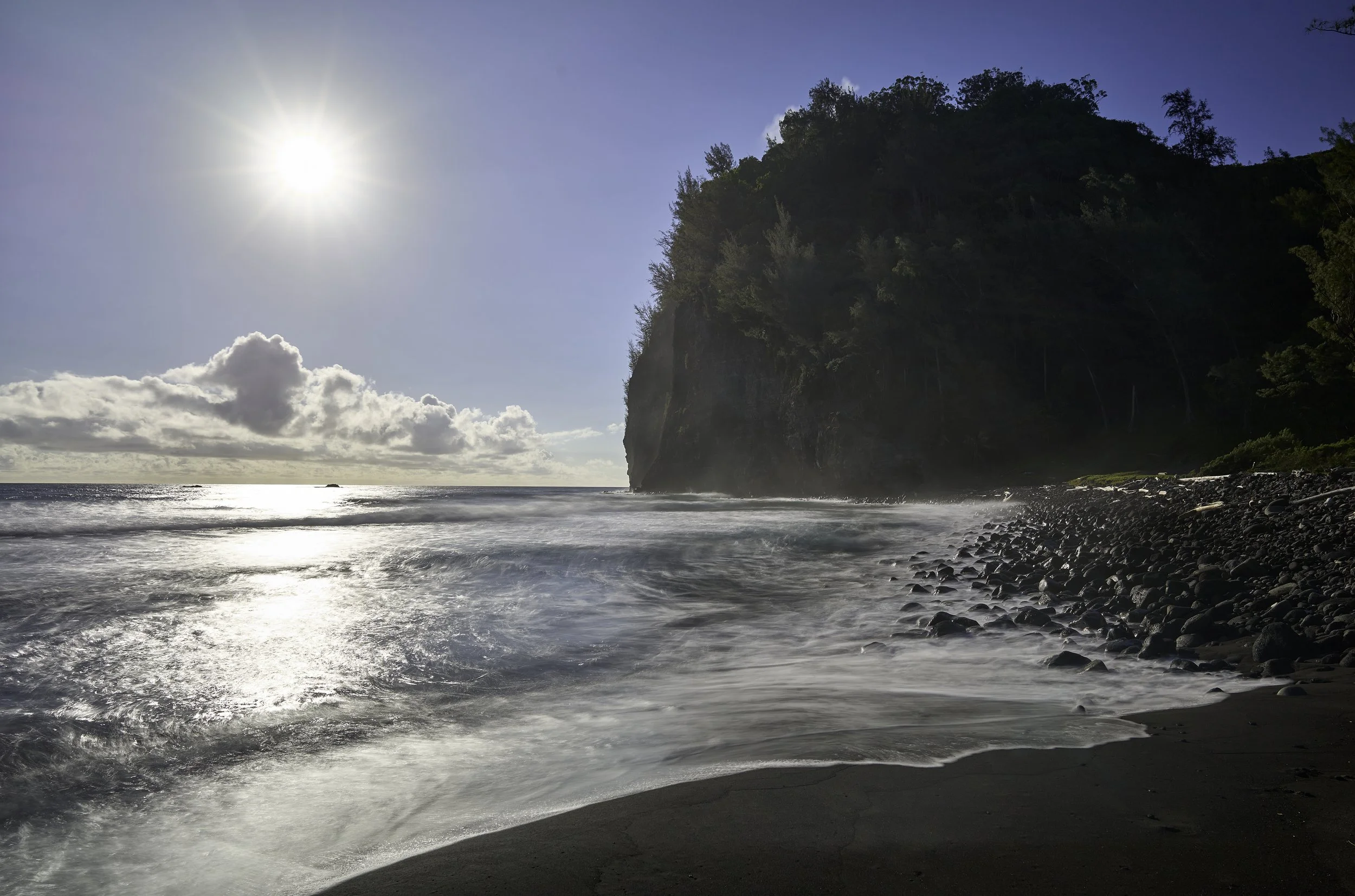 Sun shining over a Pololu Beach with waves crashing against the shore and a large tree-covered cliff on the right side of the image.