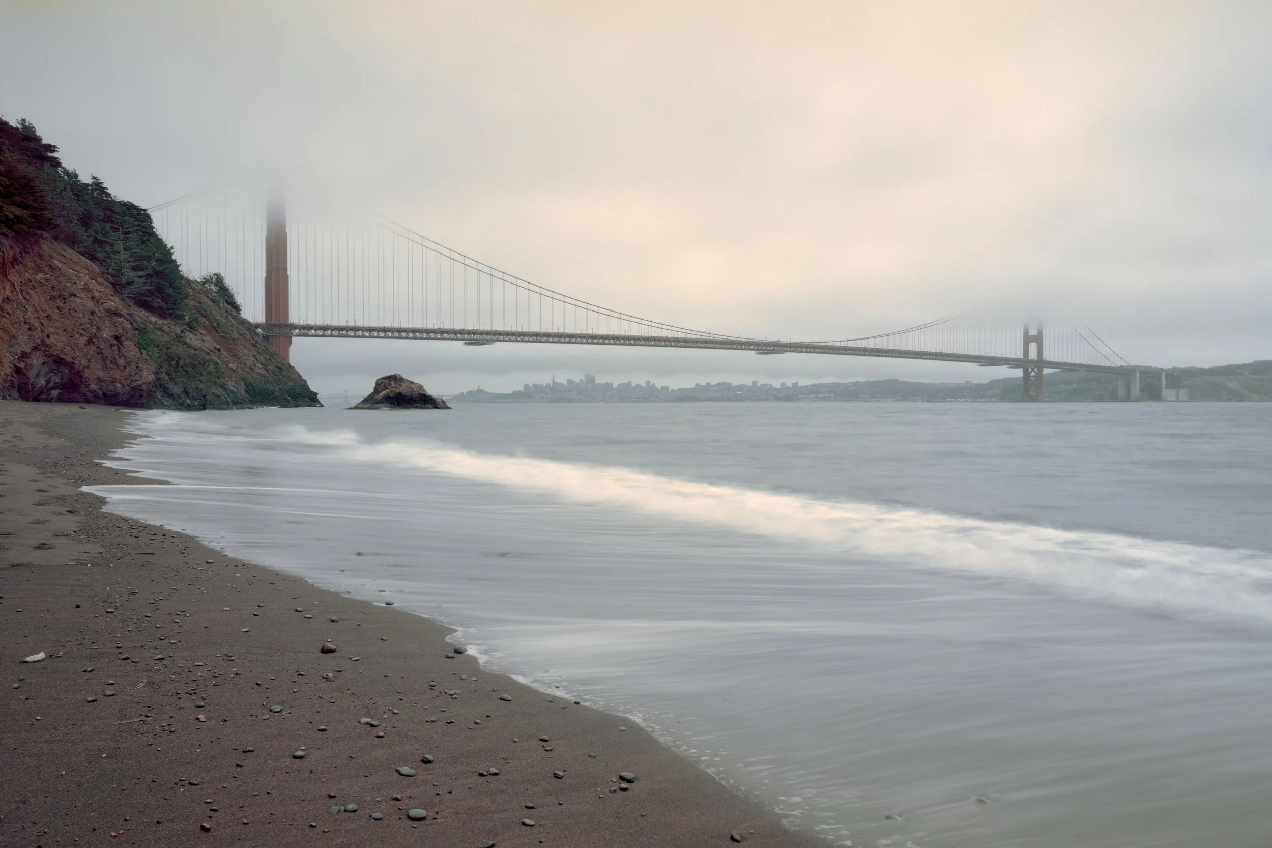 A foggy view of the Golden Gate Bridge from Kirby Cove with sand and rocks, and distant city skyline in the background.