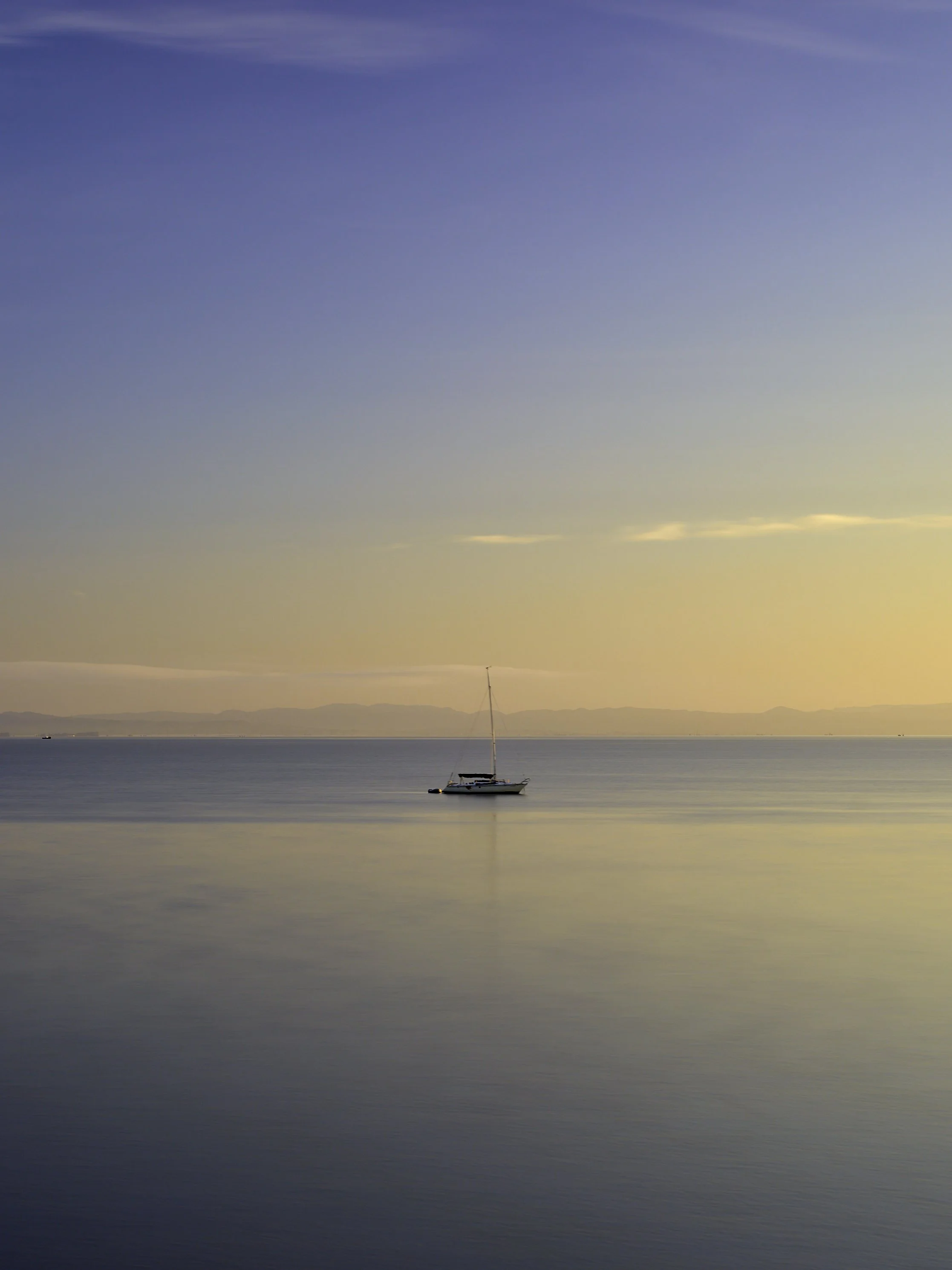 A sailboat floating on calm water at sunset with distant mountains and a clear sky. Rat Rock Cove near China Camp, San Rafael, CA.