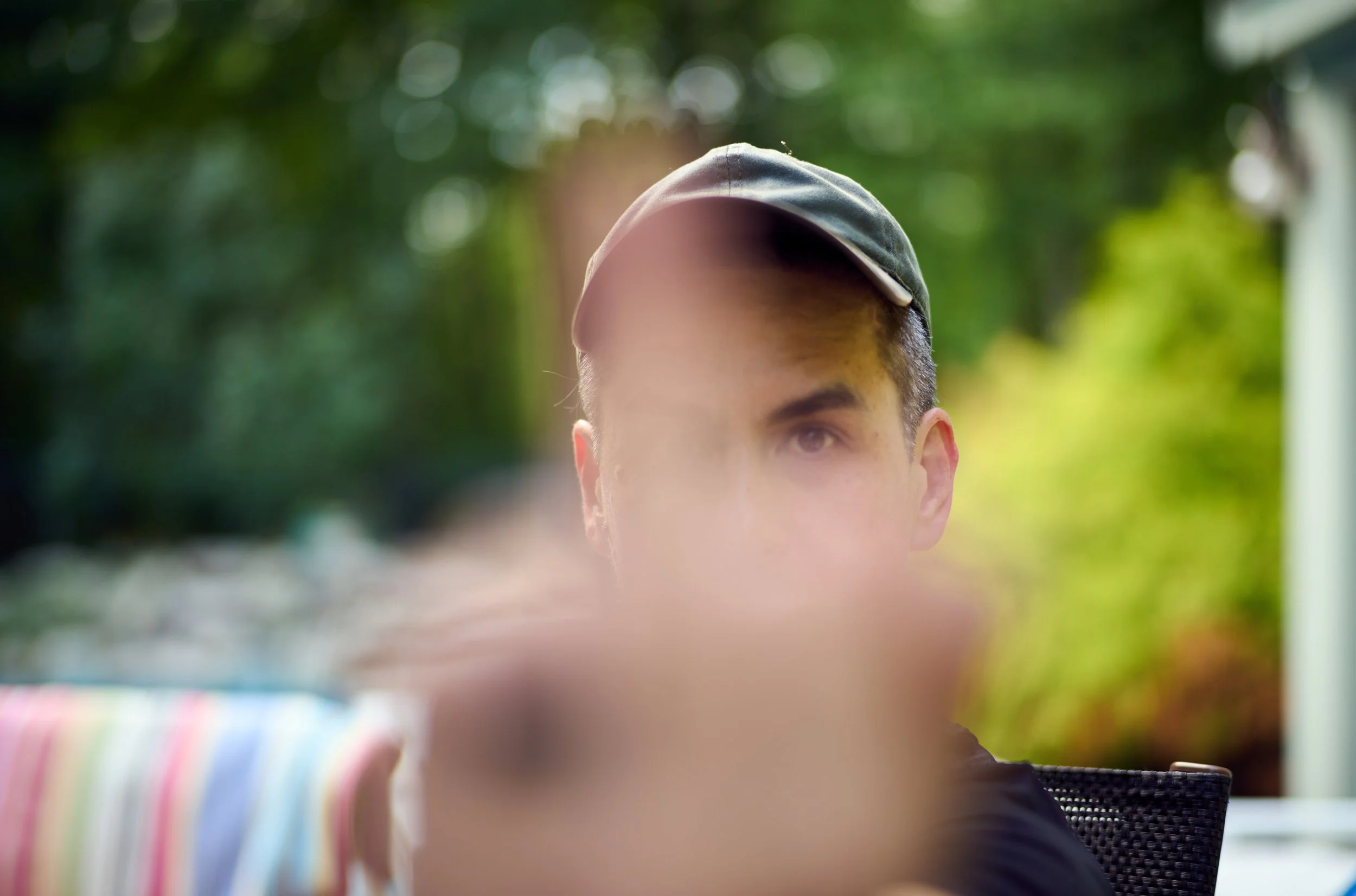 Close-up of a person's face with a baseball cap, out of focus, with blurred outdoor background and colorful striped object in foreground.