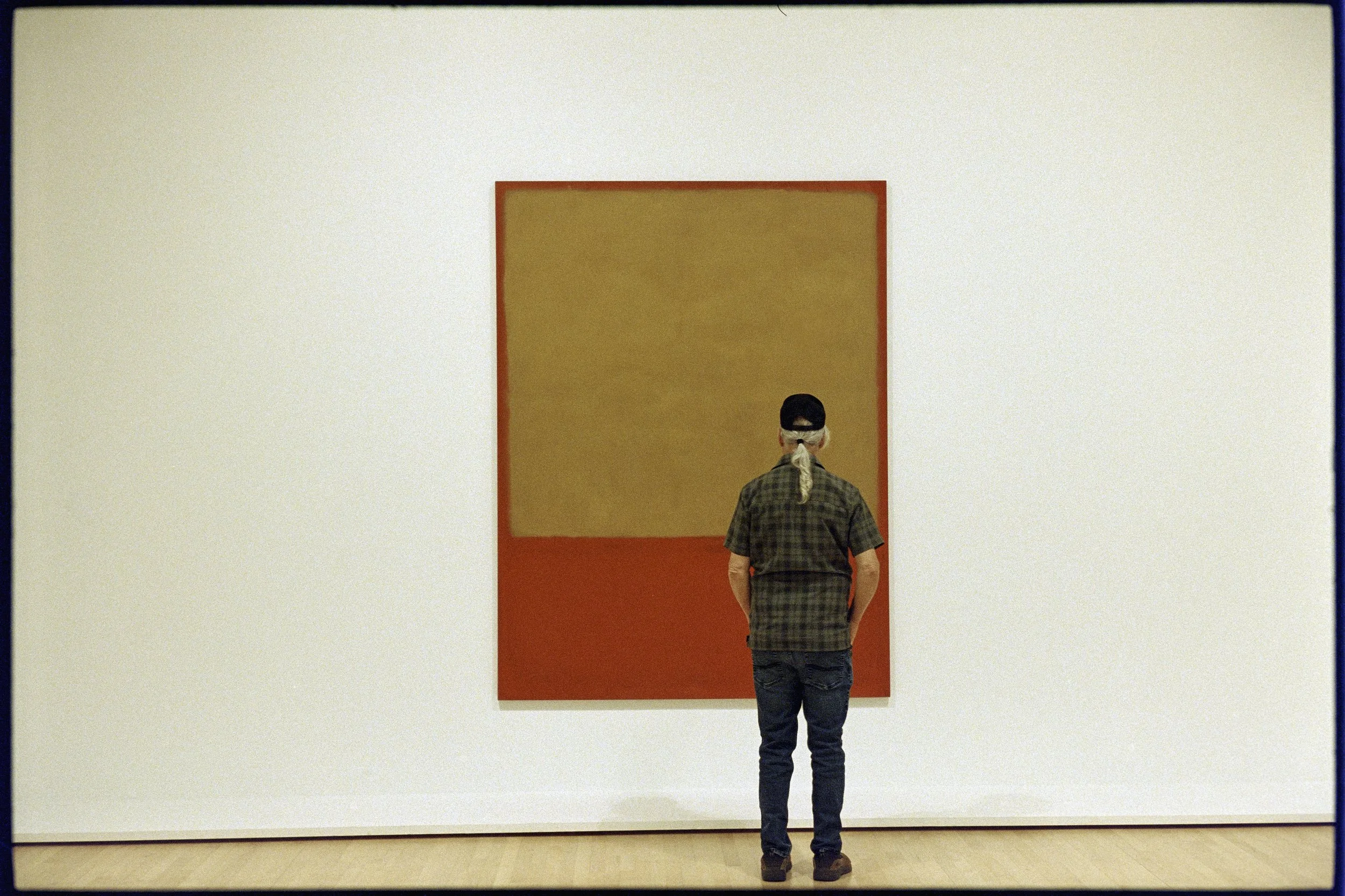 A person with long gray hair, wearing a black cap, glasses, a plaid shirt, and blue jeans, standing in front of a Mark Rothko work, Untitled 1956. SF MOMA, San Francisco, CA.