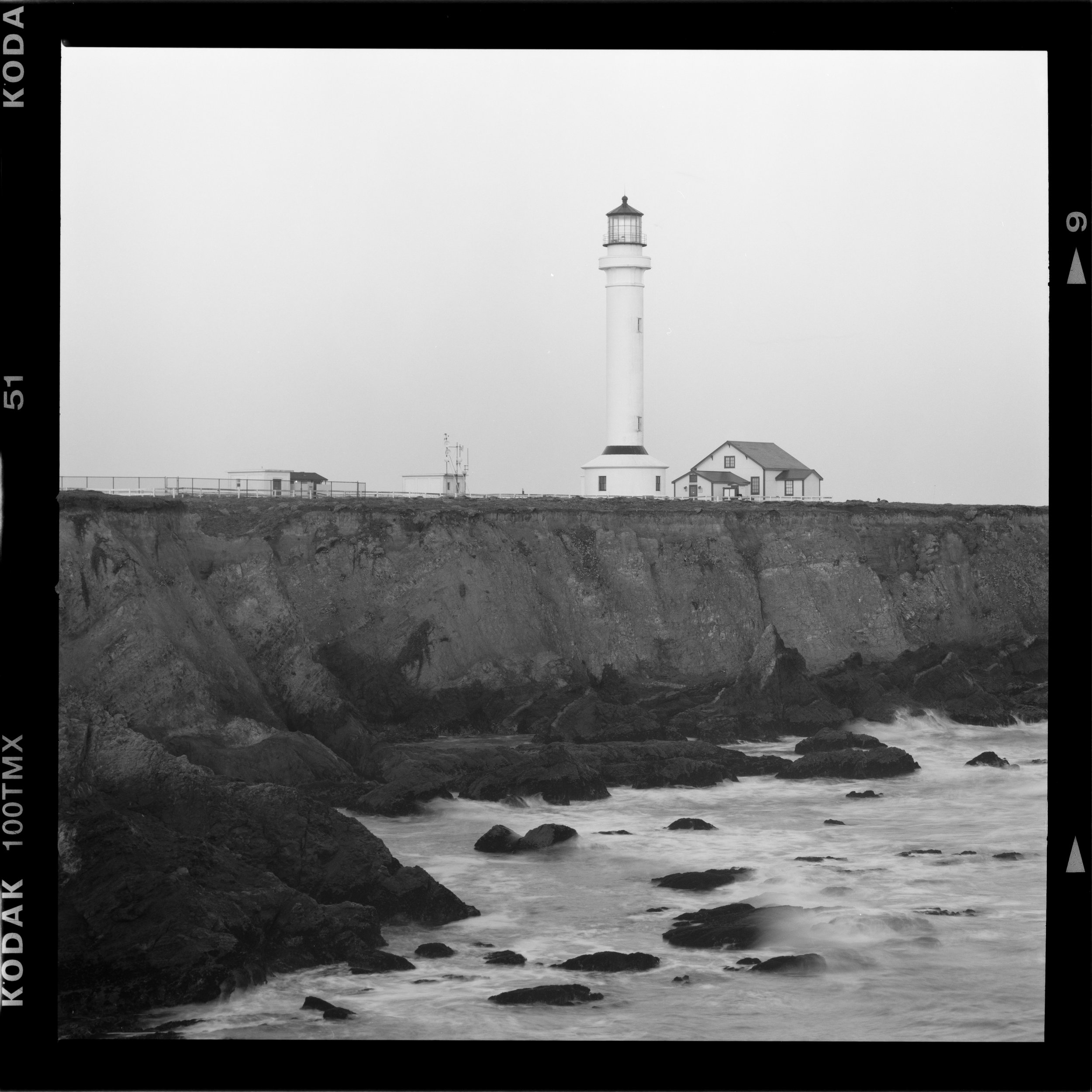 Black and white photo of a lighthouse on a coastal cliff with houses nearby and rocks in the water at the shoreline. Point Arena Lighthouse.
