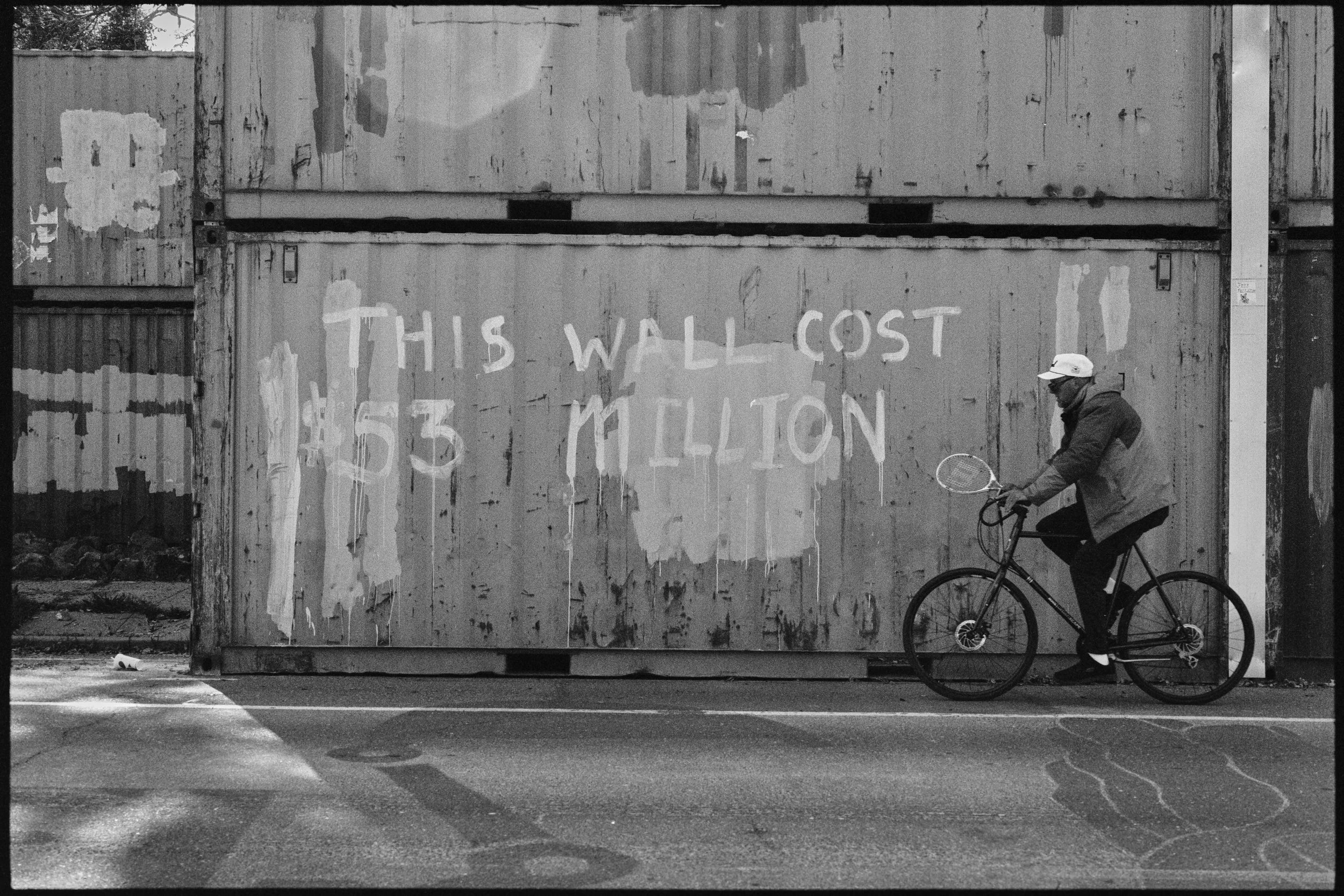A large, weathered shipping container with graffiti that reads 'This wall cost $53 million' is being walked past by a person riding a bicycle. The image is in black and white. The People's Park, Berkeley, CA.