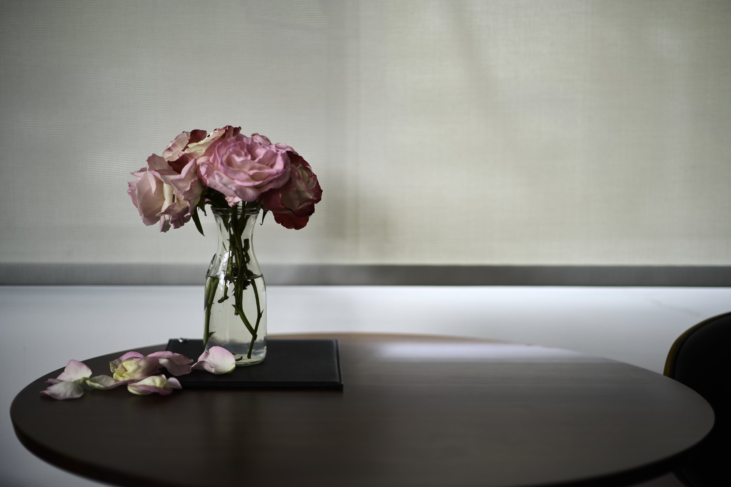 Pink and white roses in a clear glass vase on a table, with rose petals scattered around, against a plain white background.