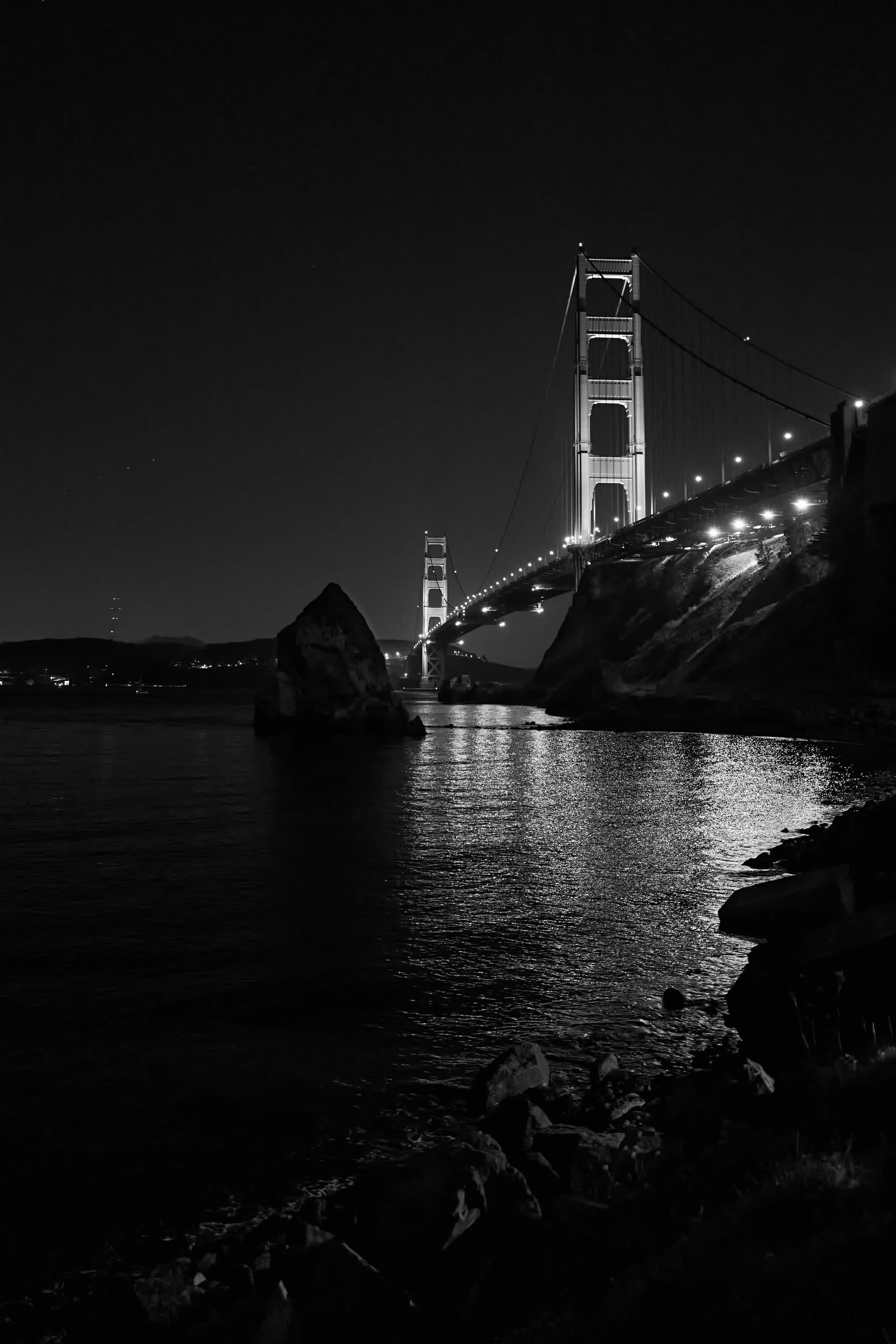 Nighttime view of the Golden Gate Bridge with illuminated towers, water reflection, and rocky shoreline in black and white.