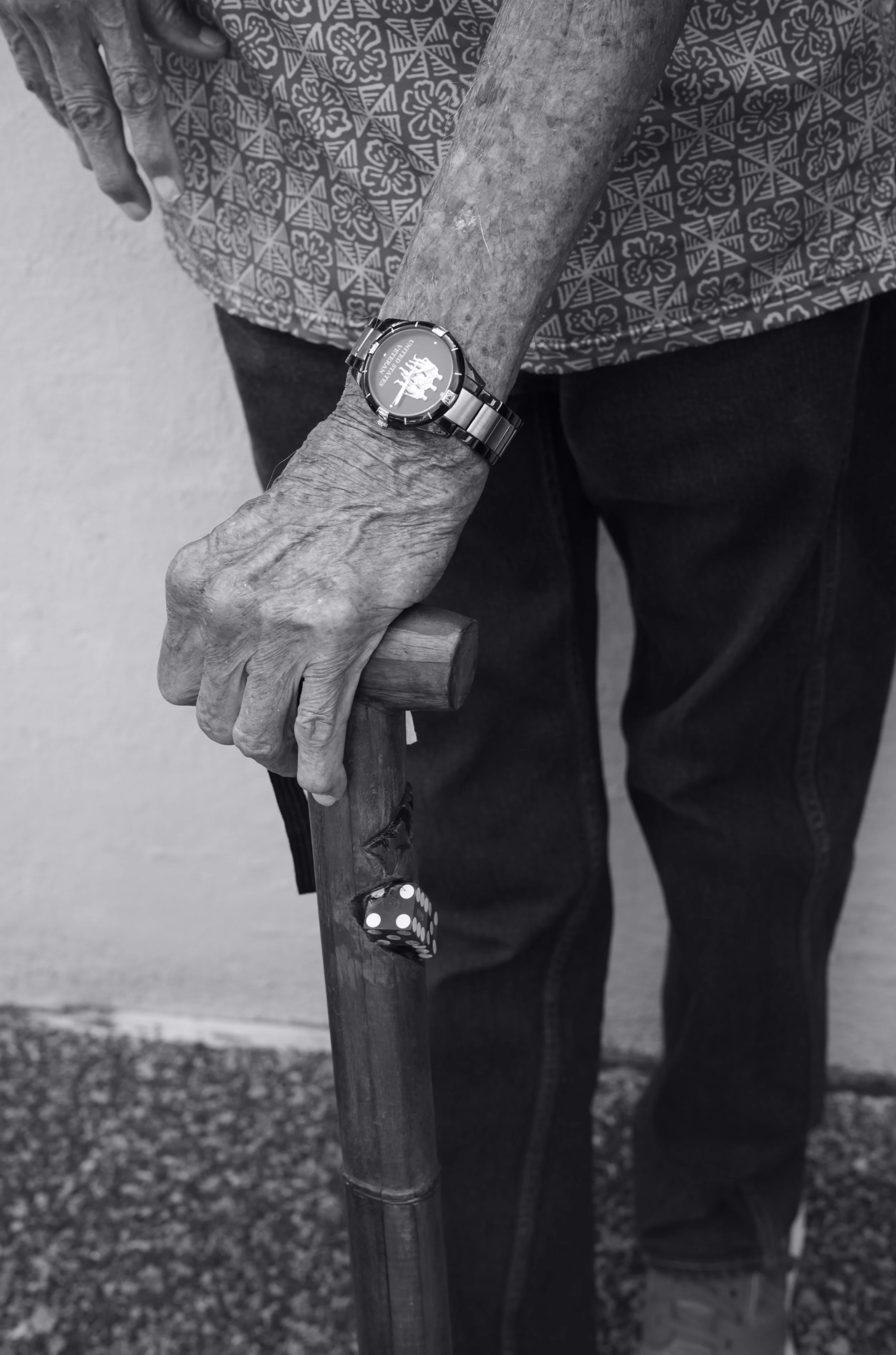 Close-up of an elderly person's hand holding a wooden cane, wearing a wristwatch, and a pair of dice embedded within the cane. The person is wearing a patterned shirt. The background shows an outdoor setting.