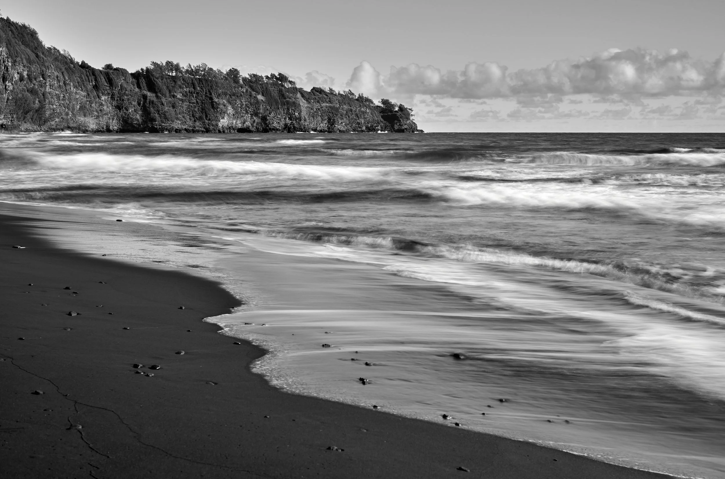 Black and white photograph of a Pololu Beach with waves crashing onto the shoreline and a rocky cliff in the background. Big Island, HI.