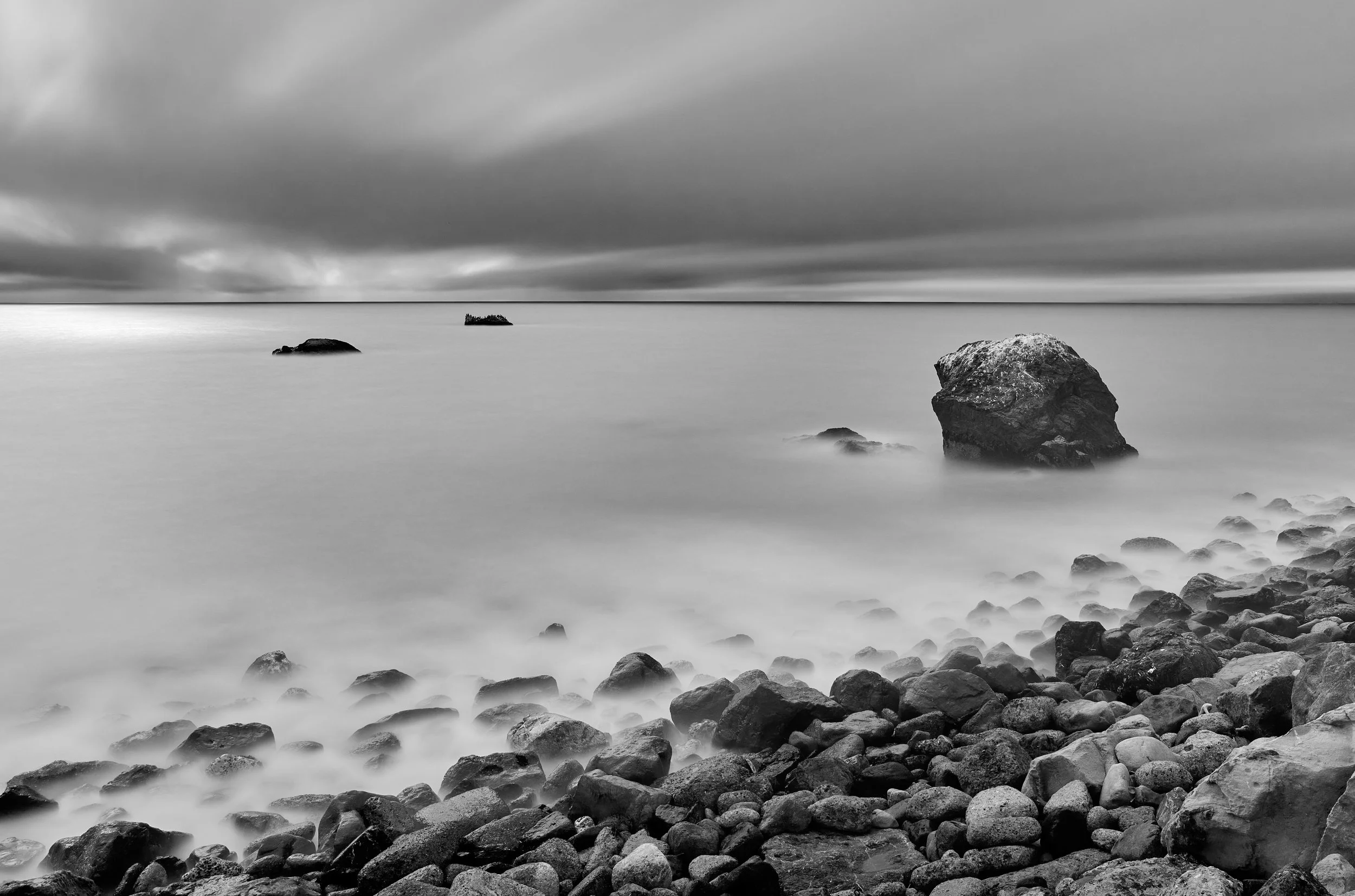 Black and white photograph of a rocky shoreline with a large rock in the foreground, smaller rocks in the water, and an overcast sky with thick clouds. Muscle Rock Park, Daly City, CA.