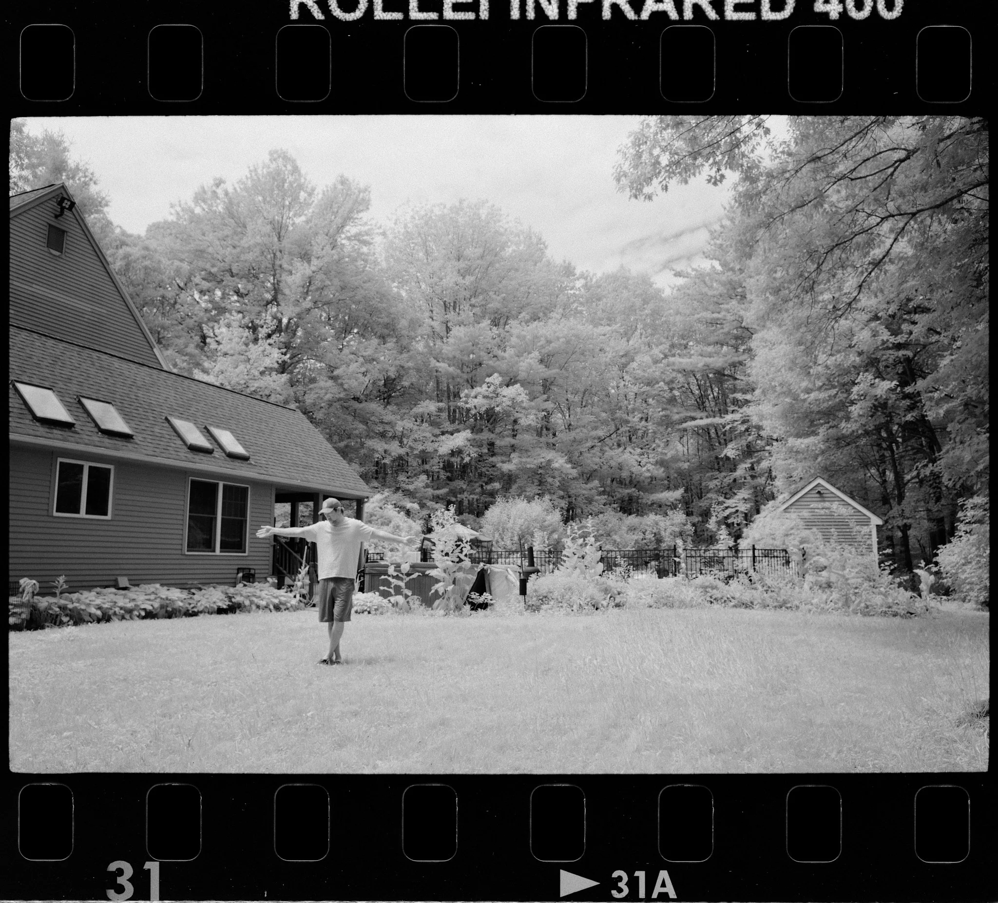 A black and white image showing a backyard with trees, a house with skylights, and a person standing on the grass with arms outstretched. Infrared sensitive film.