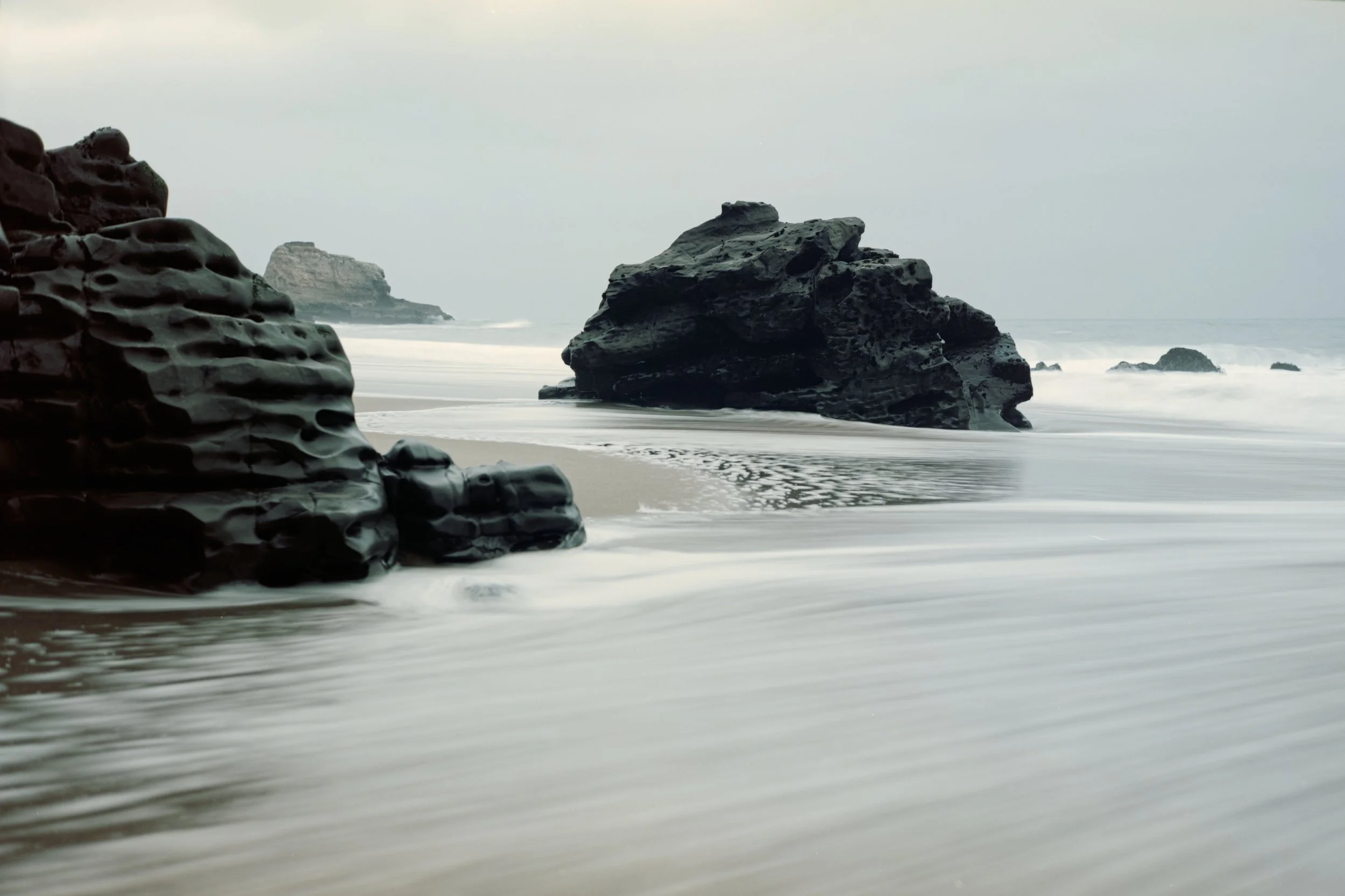 Overcast day at Panther Beach with large dark rocks in the foreground and distant cliffs. Santa Cruz, CA.