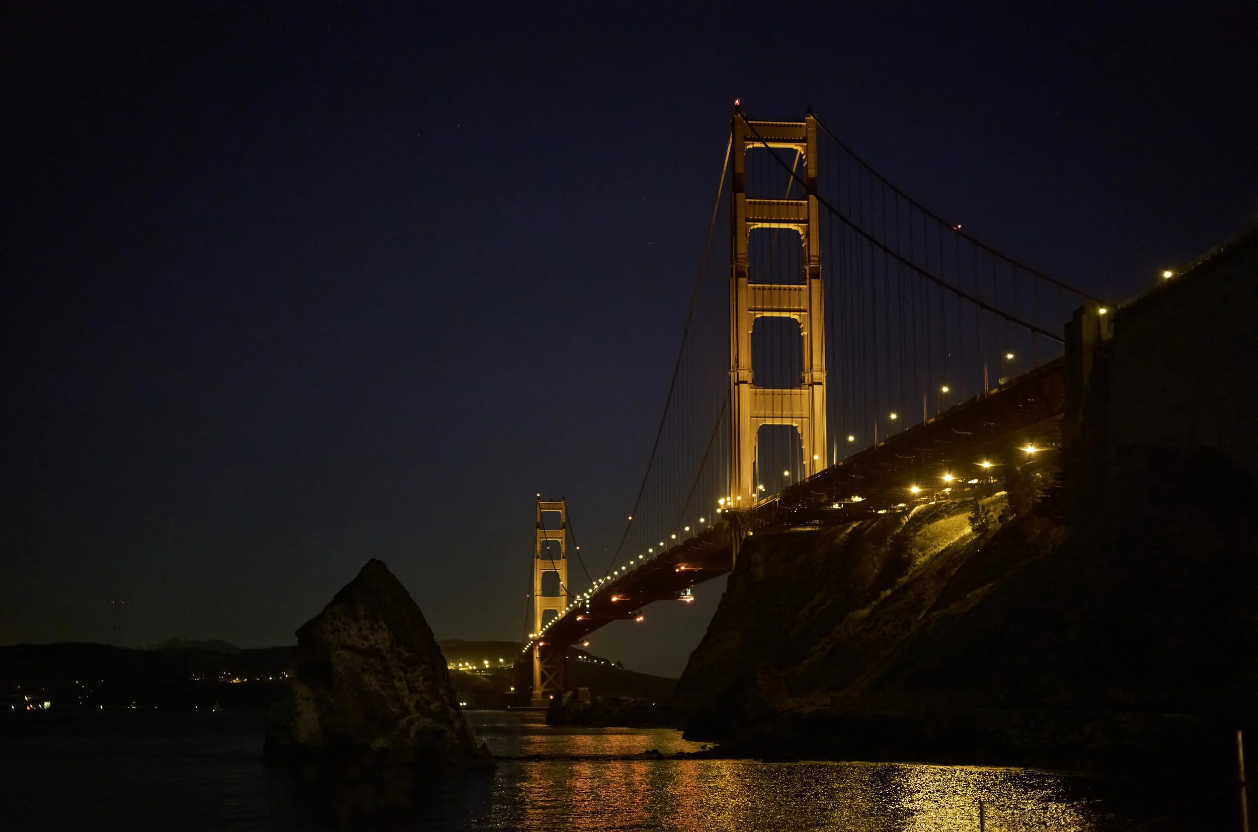 Night view of the Golden Gate Bridge illuminated, with reflections on the water, and a dark sky with some stars.