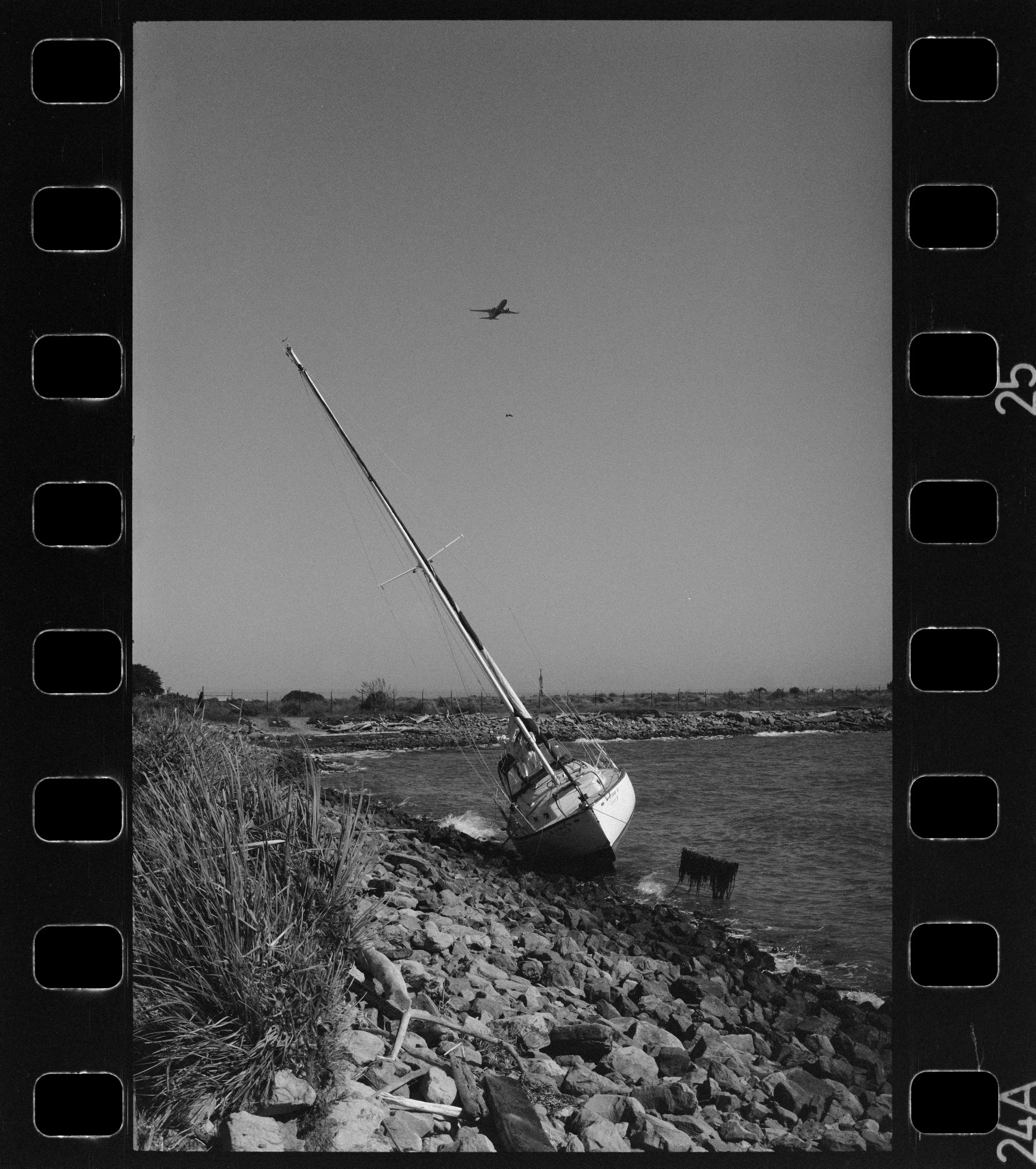 Black and white photograph of a boat beached on a rocky shoreline of the bay, with a tall mast leaning at an angle and a small airplane flying high in the sky above.