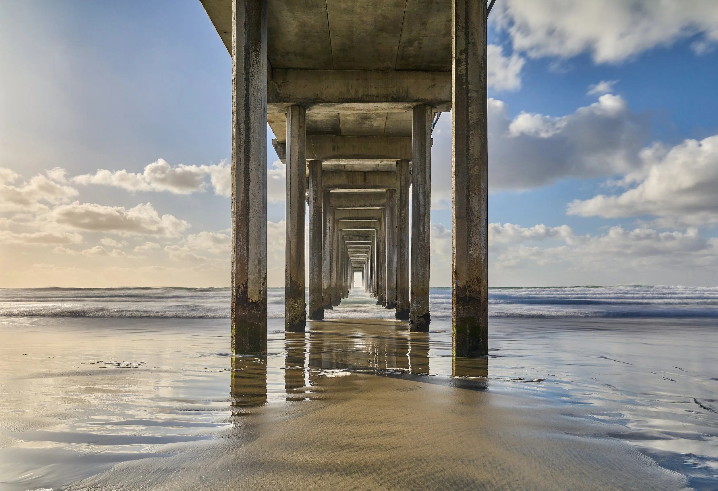 View underneath UCSD Scripps Pier on the beach with wooden support beams extending into the ocean. The sky is partly cloudy with sunlight illuminating the scene. La Jolla, CA.