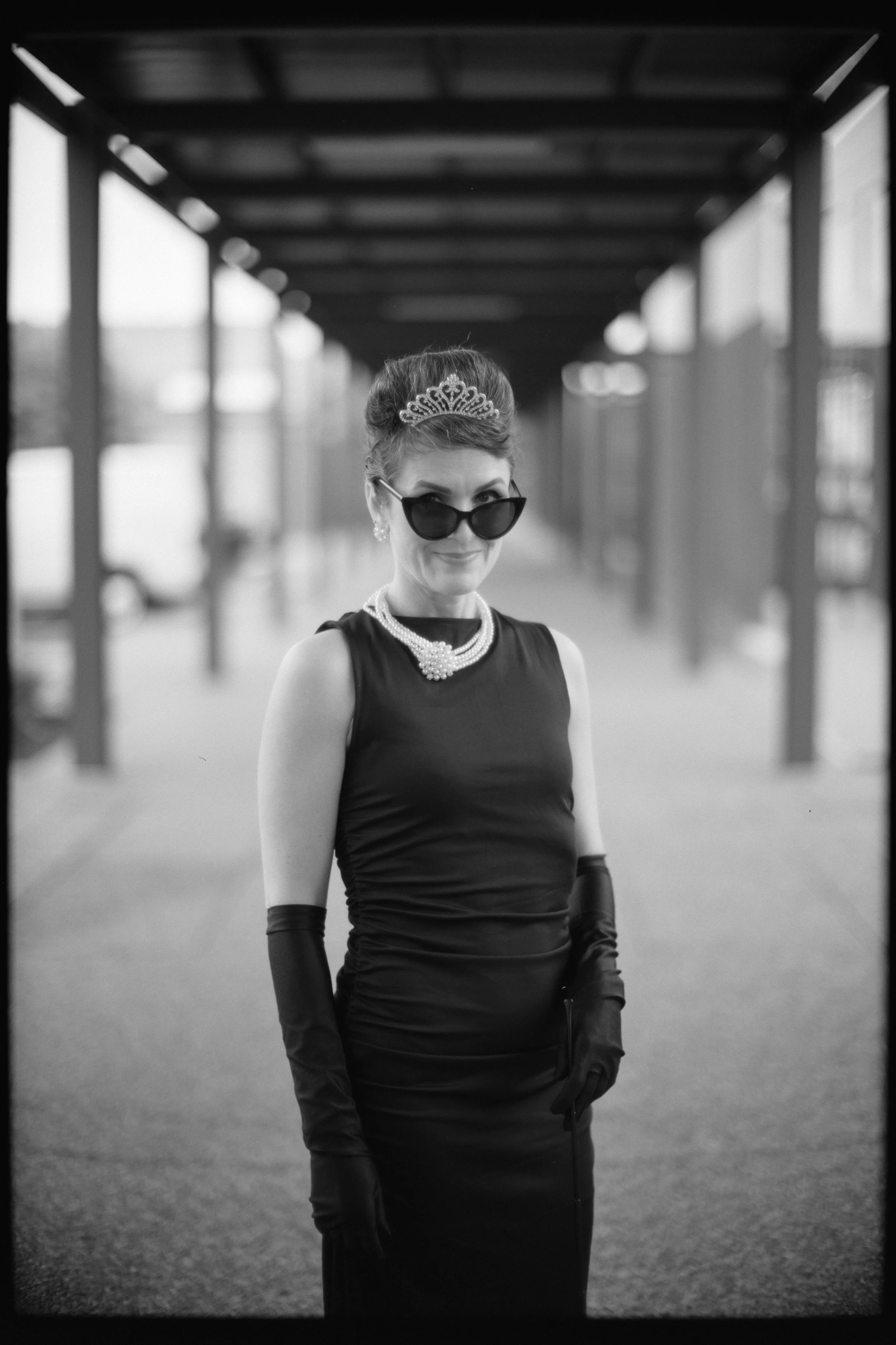 A woman dressed as a glamorous 1950s style character, wearing a tiara, sunglasses, pearl necklace, sleeveless dress, and elegant gloves, stands under an outdoor structure with a blurred background.
