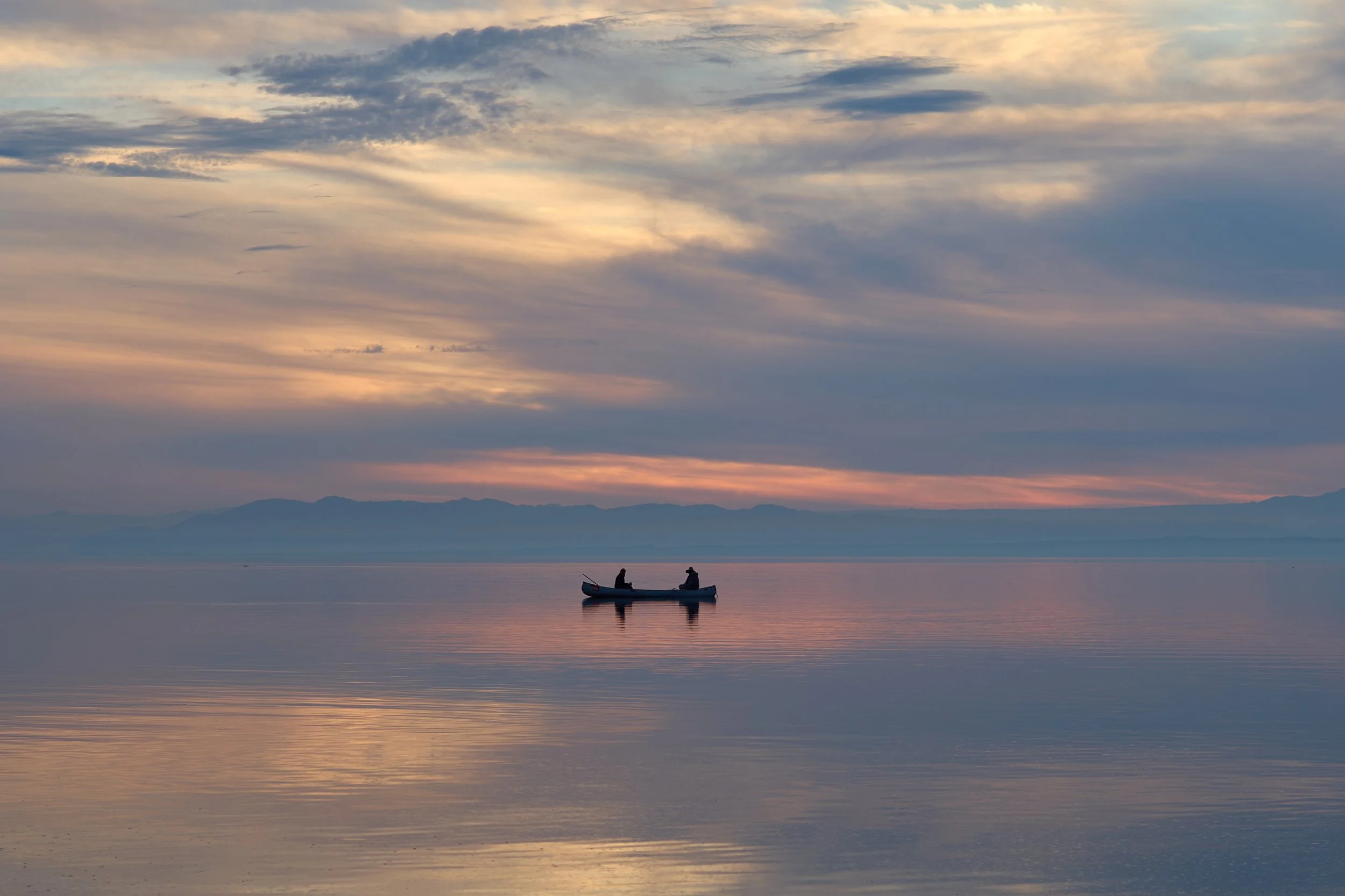 Two people in a small boat on calm water during sunset, with colorful clouds in the sky and mountains in the distance. Bombay Beach, Salton Sea, CA.