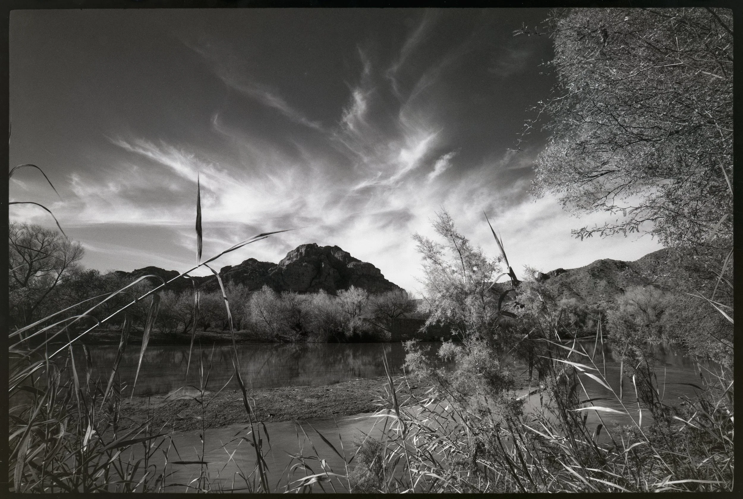 A black and white landscape photograph showing a river in the foreground with tall grasses along its banks, a rocky mountain in the middle distance, and trees on either side. The sky above has wispy clouds. Salt River, AZ.