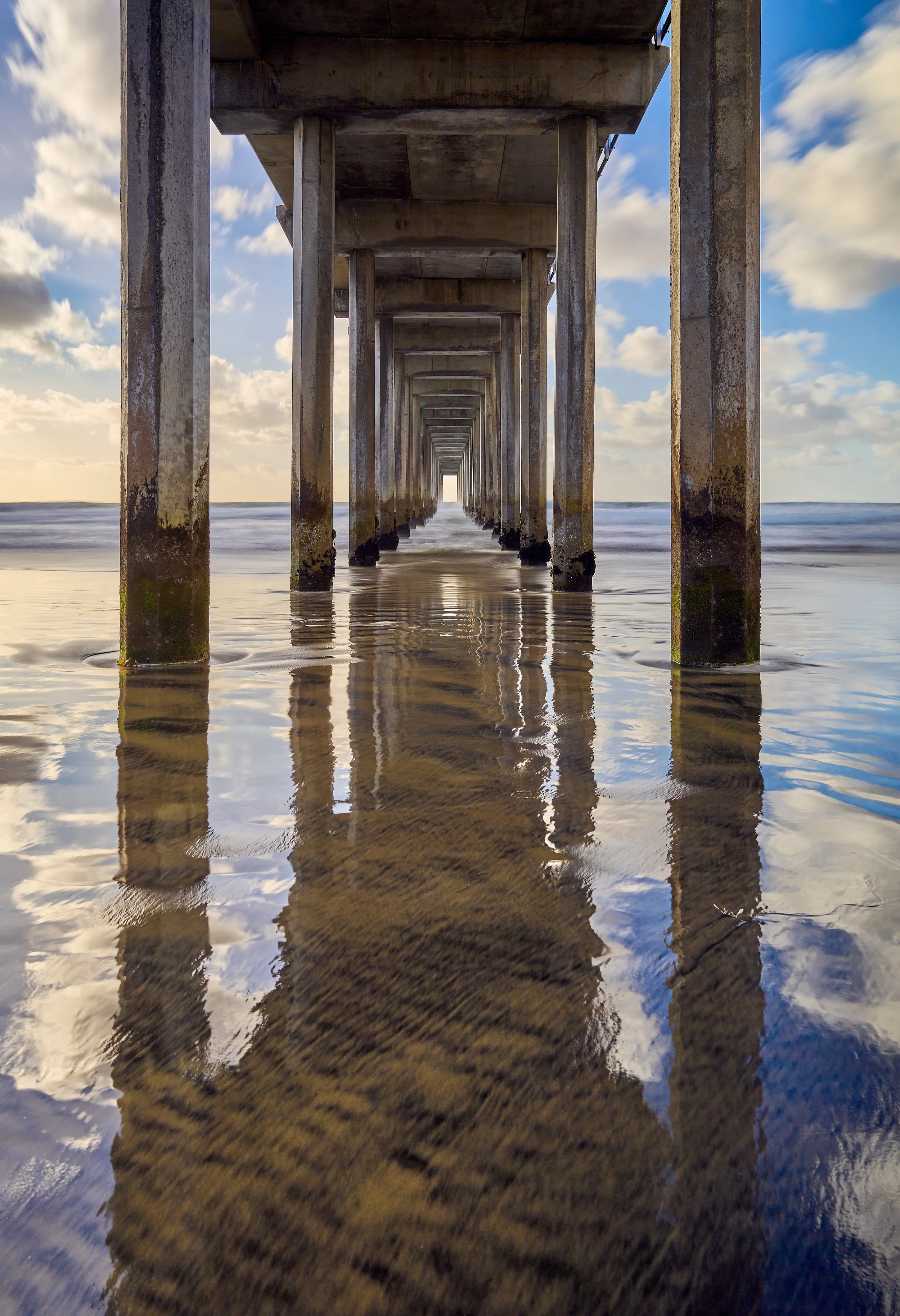 View under UCSD Scripps Pier at the beach with concrete pillars reflected in wet sand, clouds in the sky, and ocean waves in the background. La Jolla, CA.