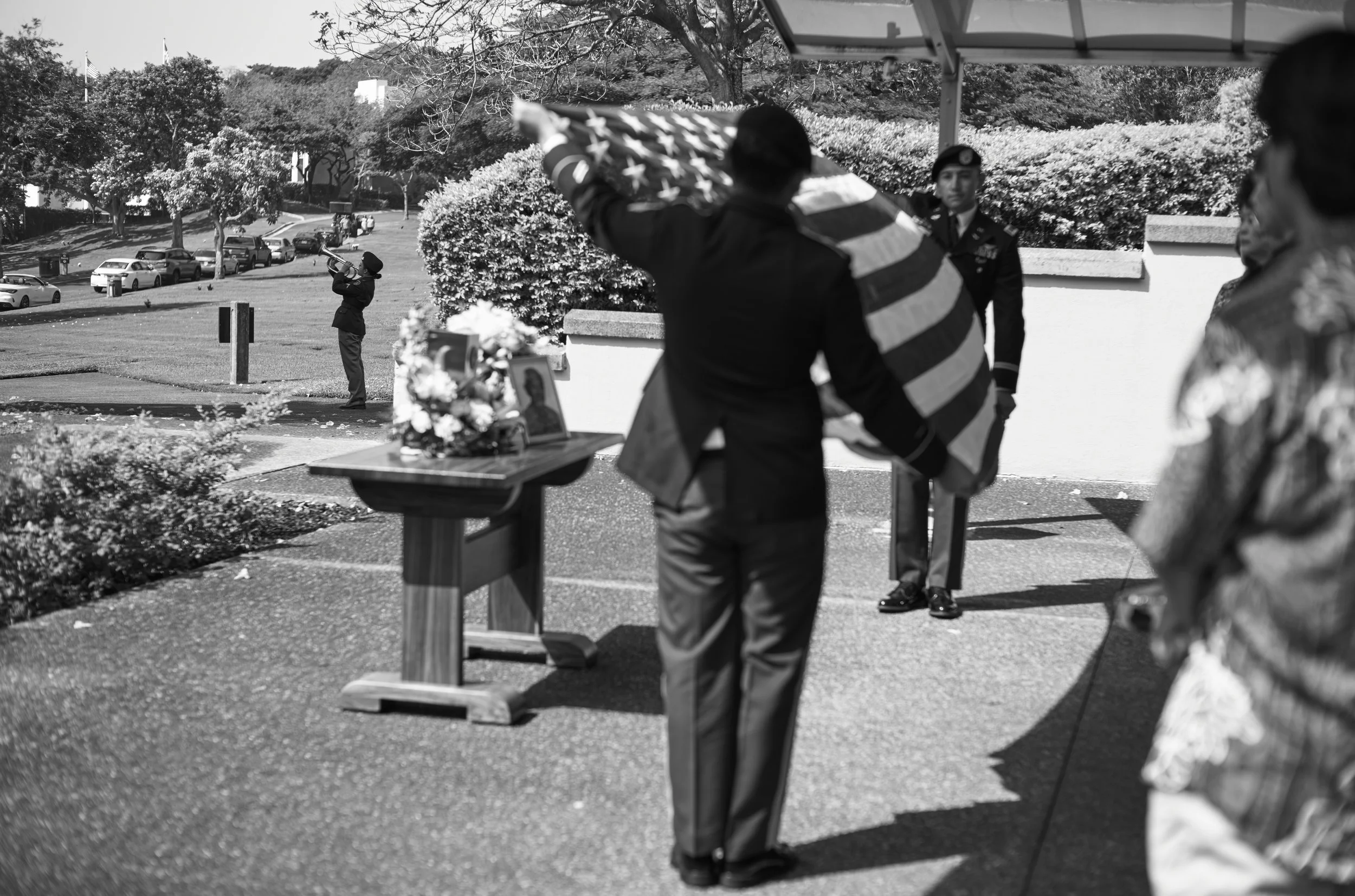 A black and white photo of a memorial ceremony outdoors with military personnel and civilians. A person in uniform performs a trumpet salute, and a man holds a folded American flag. There is a table with flowers and a photo. Several cars are parked o